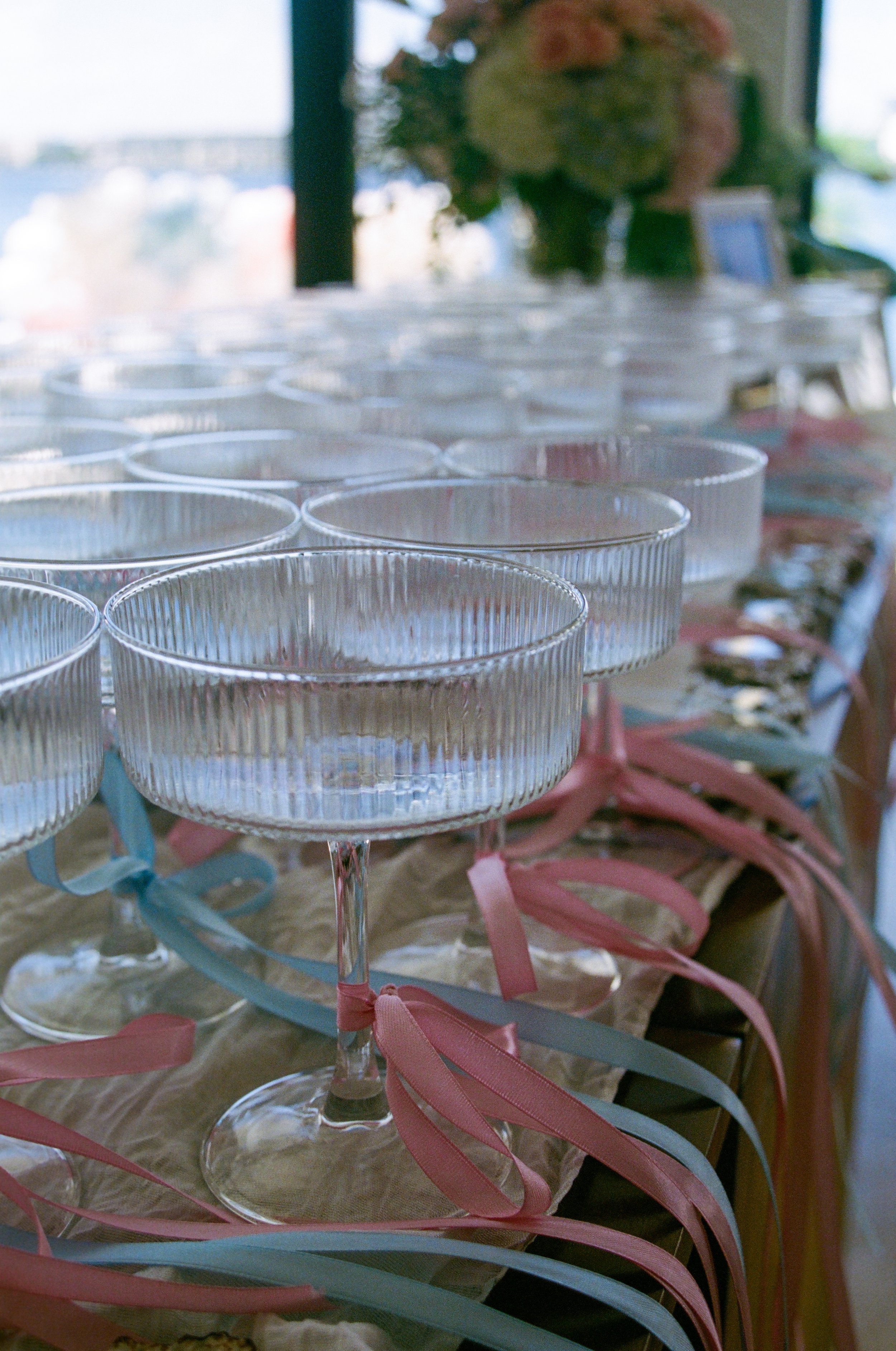Several empty clear glass dessert cups with pastel-colored ribbons tied around the stems, arranged in rows on a table, with colorful decorations in the background.