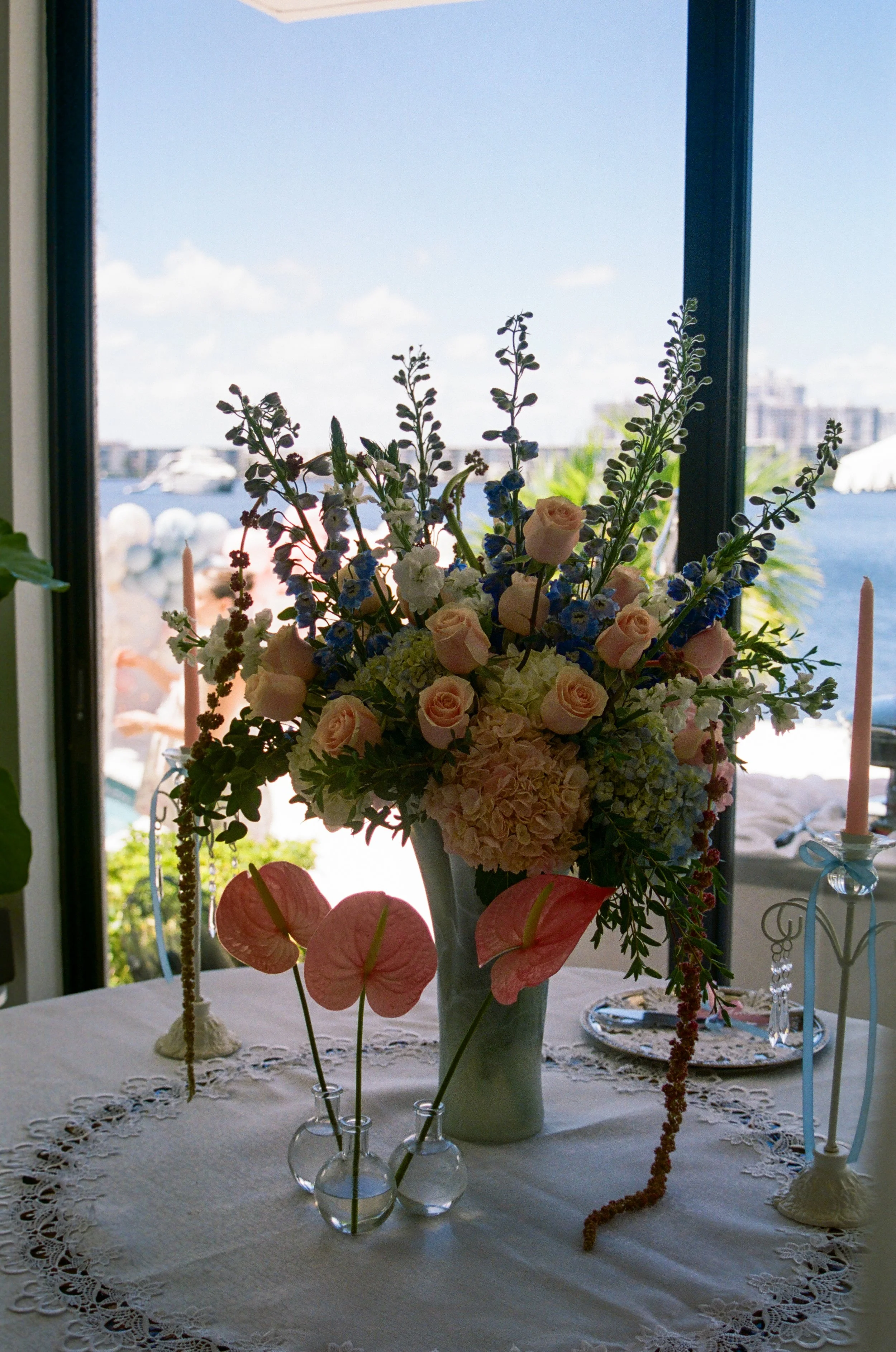 A tall floral arrangement on a table with a white lace tablecloth, featuring pink roses, white and blue hydrangeas, and other flowers in a white vase. In the background, a window shows a view of a body of water with boats and a city skyline.