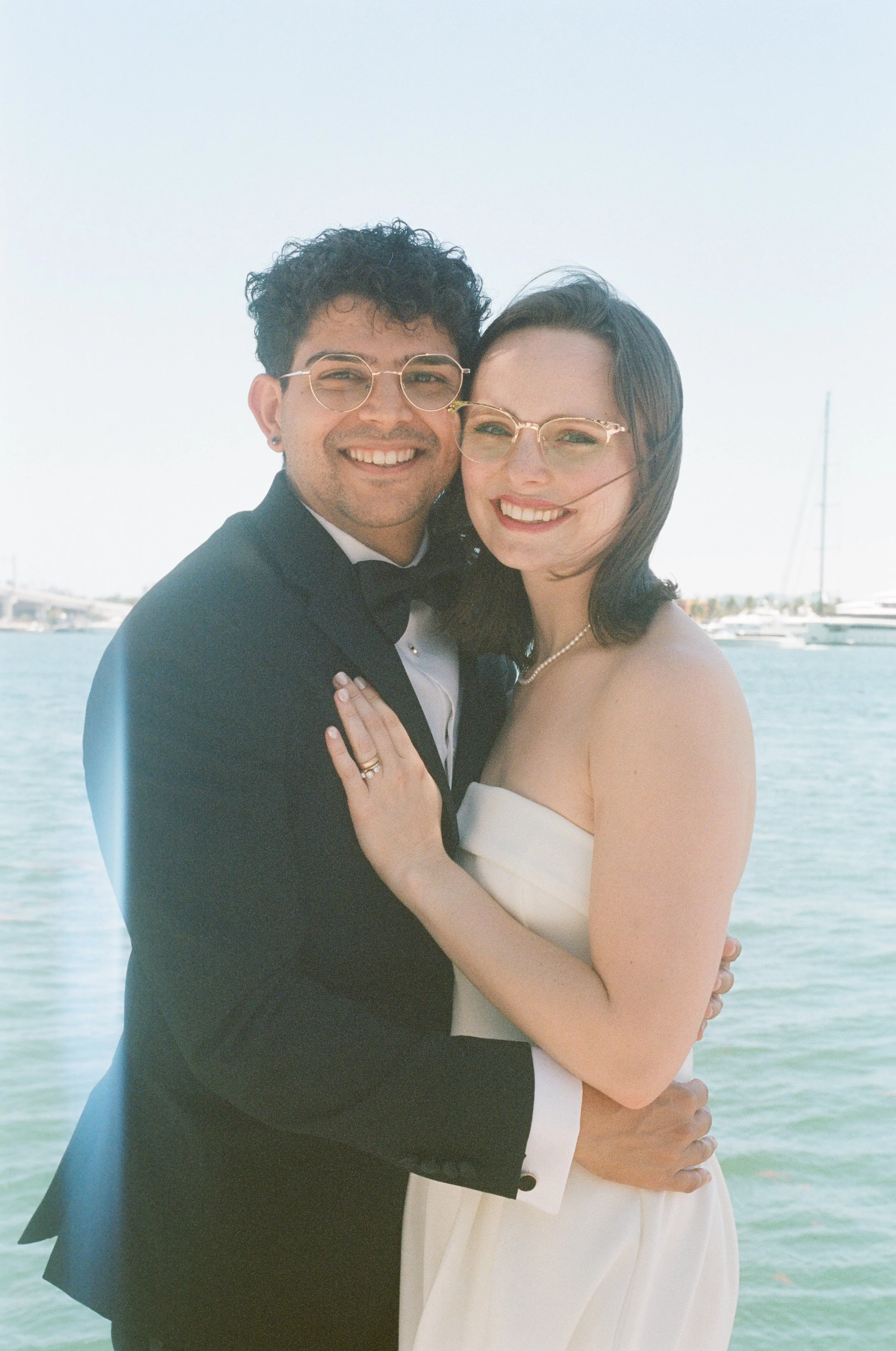 A happily married couple, dressed in formal wedding attire, embracing by a body of water with boats and a dock in the background during daytime.