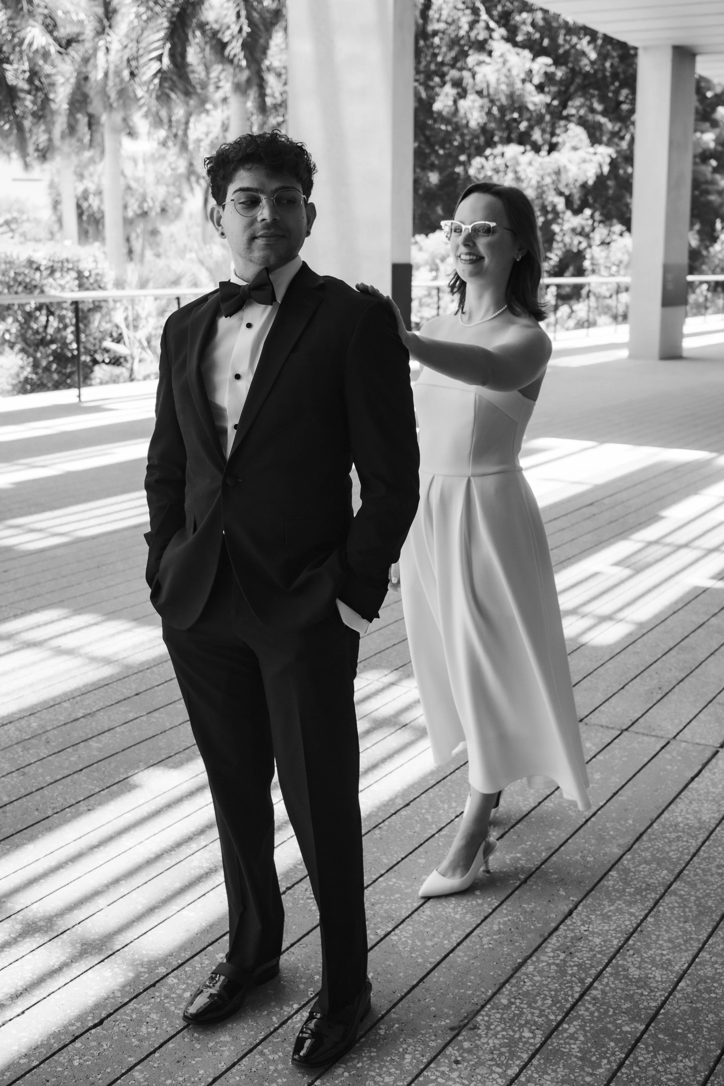 A woman in a white dress with glasses and pearl necklace is smiling and making a playful gesture with her hand on a man's shoulder, who is dressed in a tuxedo, on a wooden deck outside with trees in the background.