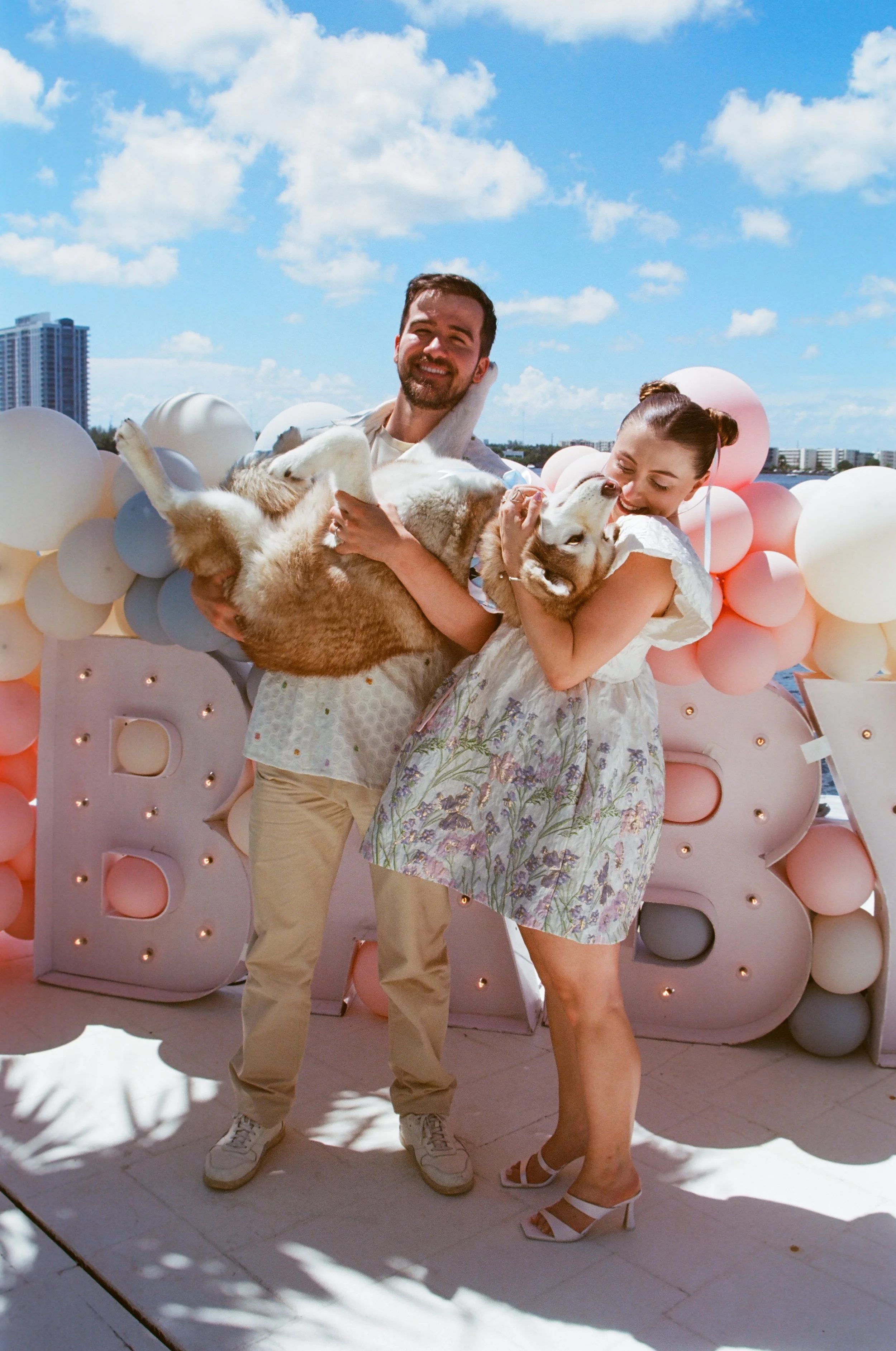 A happy couple holding two dogs in front of a decorated backdrop with balloons and a large illuminated letter 'B.' The man is wearing a white shirt and beige pants, and the woman is in a floral dress with white heels. The background shows a cityscape