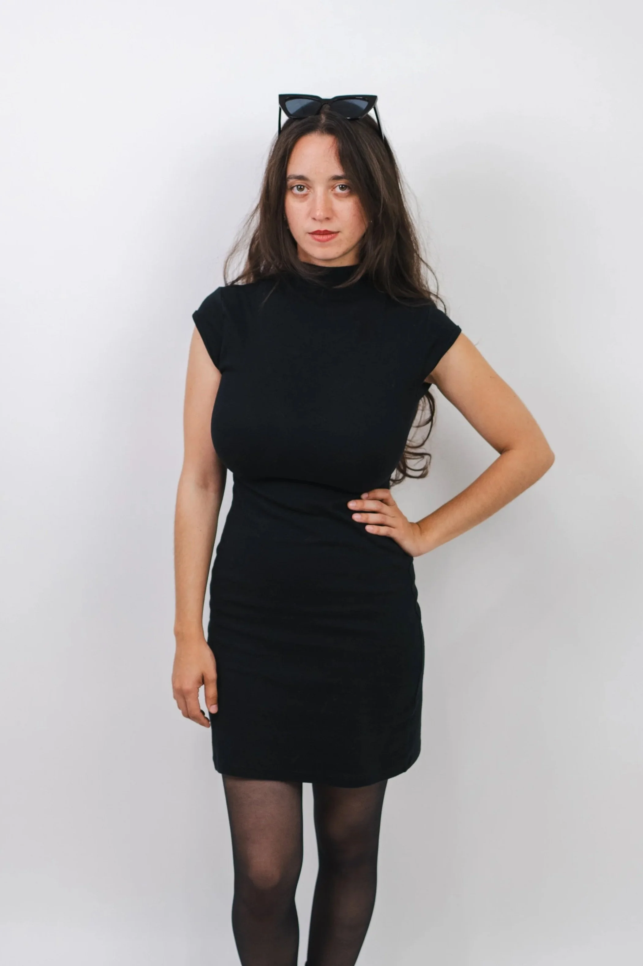 Photographer Leeanne Drucker standing with long dark hair wearing a black dress, sheer black tights, and sunglasses on her head standing against a plain white backdrop.