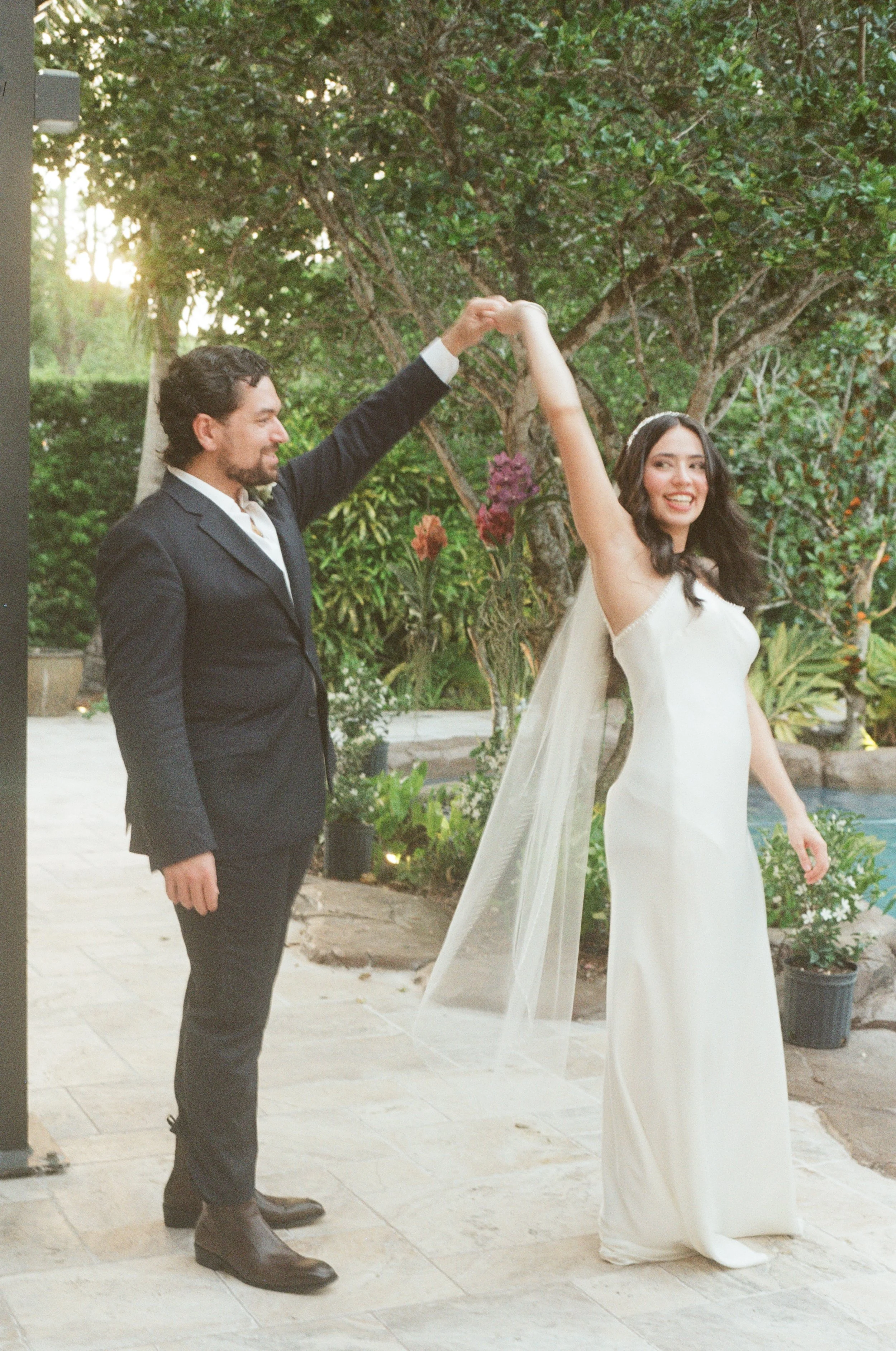 A man and woman dancing outdoors during a wedding ceremony, with the man in a black suit and the woman in a white wedding dress and veil, surrounded by greenery and flowers.