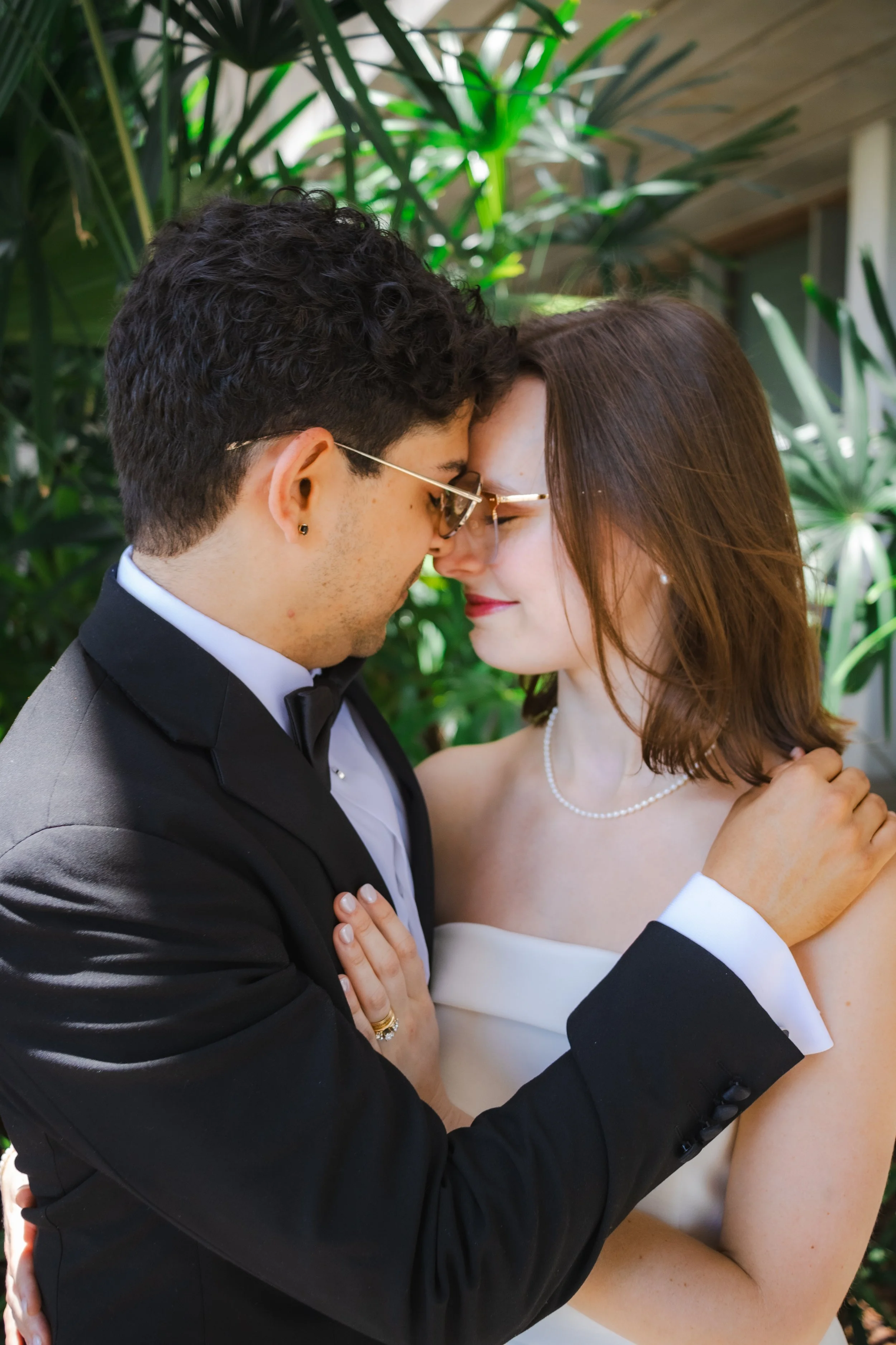A couple, dressed in formal attire, is standing close with their foreheads touching and eyes closed, surrounded by green plants.