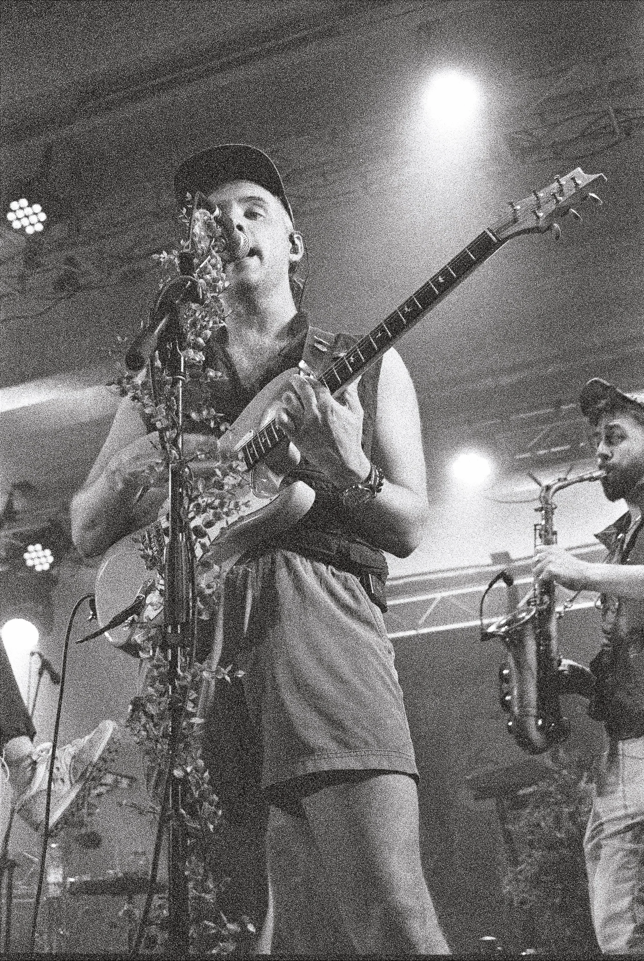 A musician playing guitar in a dimly lit venue with spotlights and other band members visible in the background.