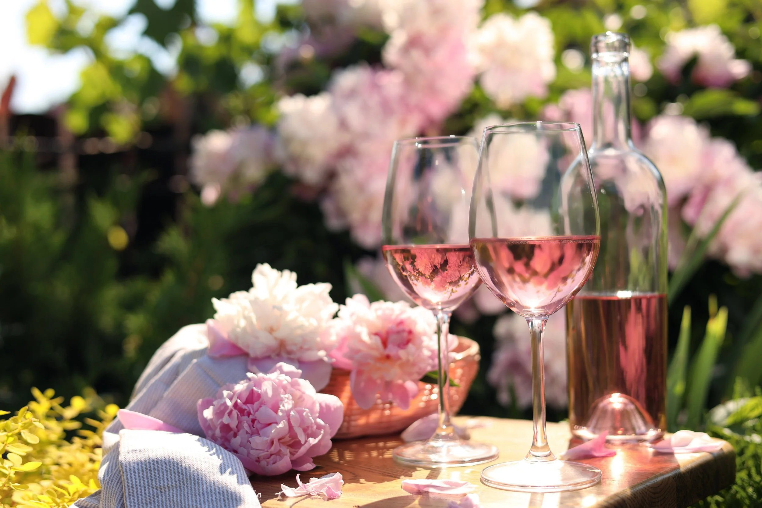 Two glasses of pink wine, a glass bottle, and a basket of pink and white flowers on a wooden table outdoors with pink flowers and green foliage in the background.