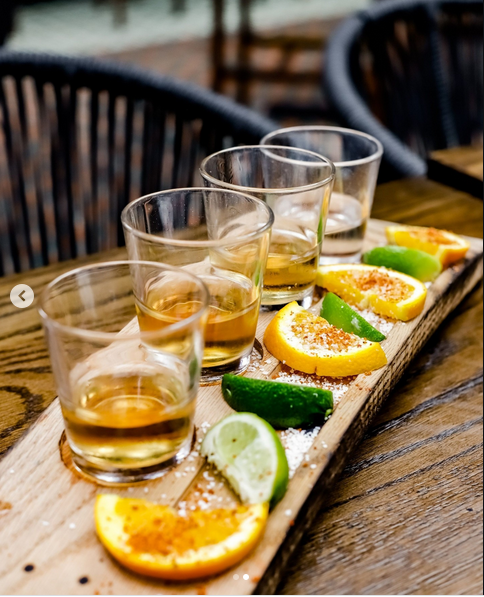 Four shot glasses filled with amber-colored liquor, alongside slices of lime and lemon on a wooden serving board, garnished with salt and sugar. The setup is on a wooden table outdoors.