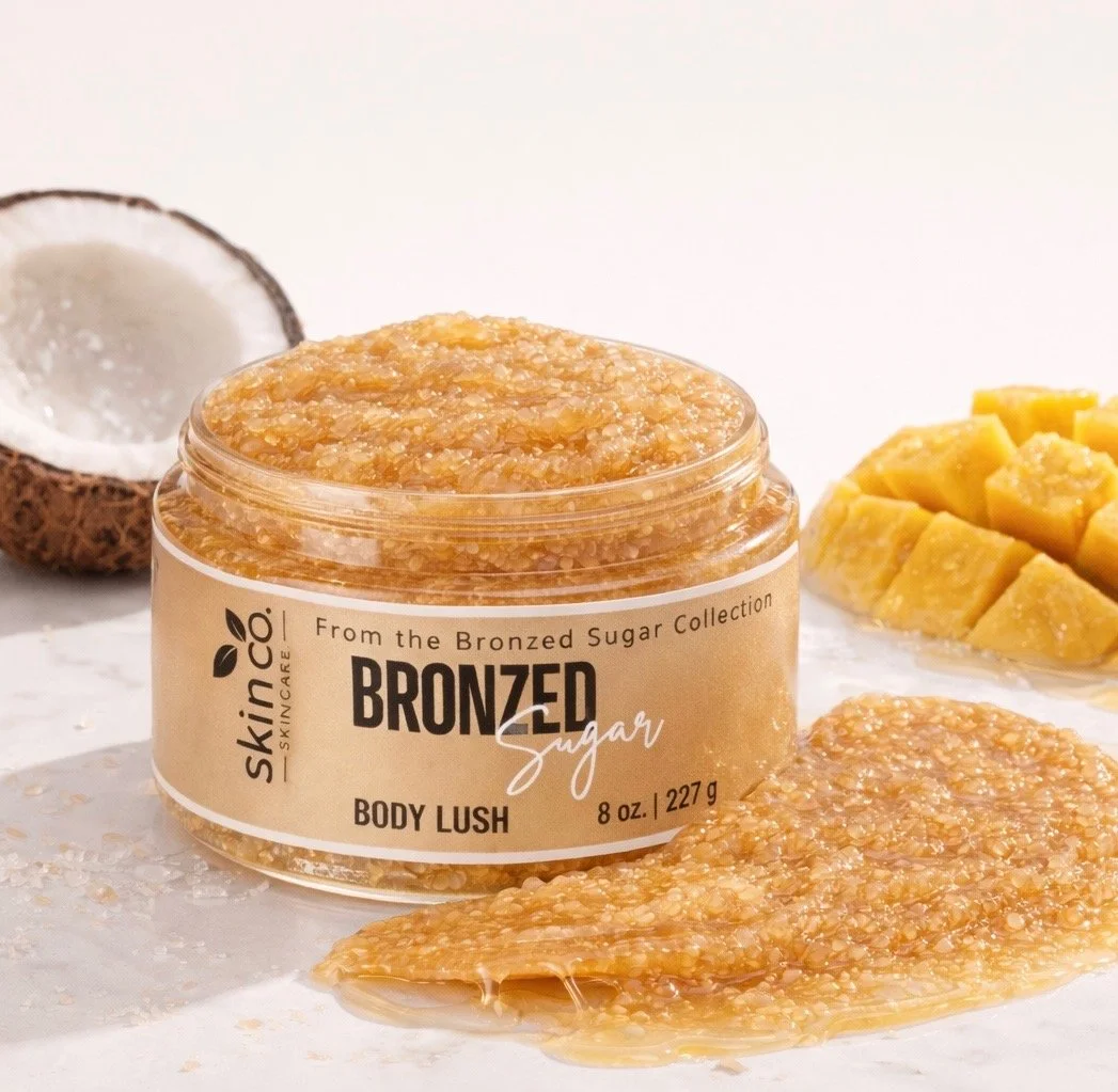 A jar of Skin&Co Bronzed Sugar Body Lush with sugar scrub spilling onto a white surface, surrounded by half a coconut and pineapple chunks.
