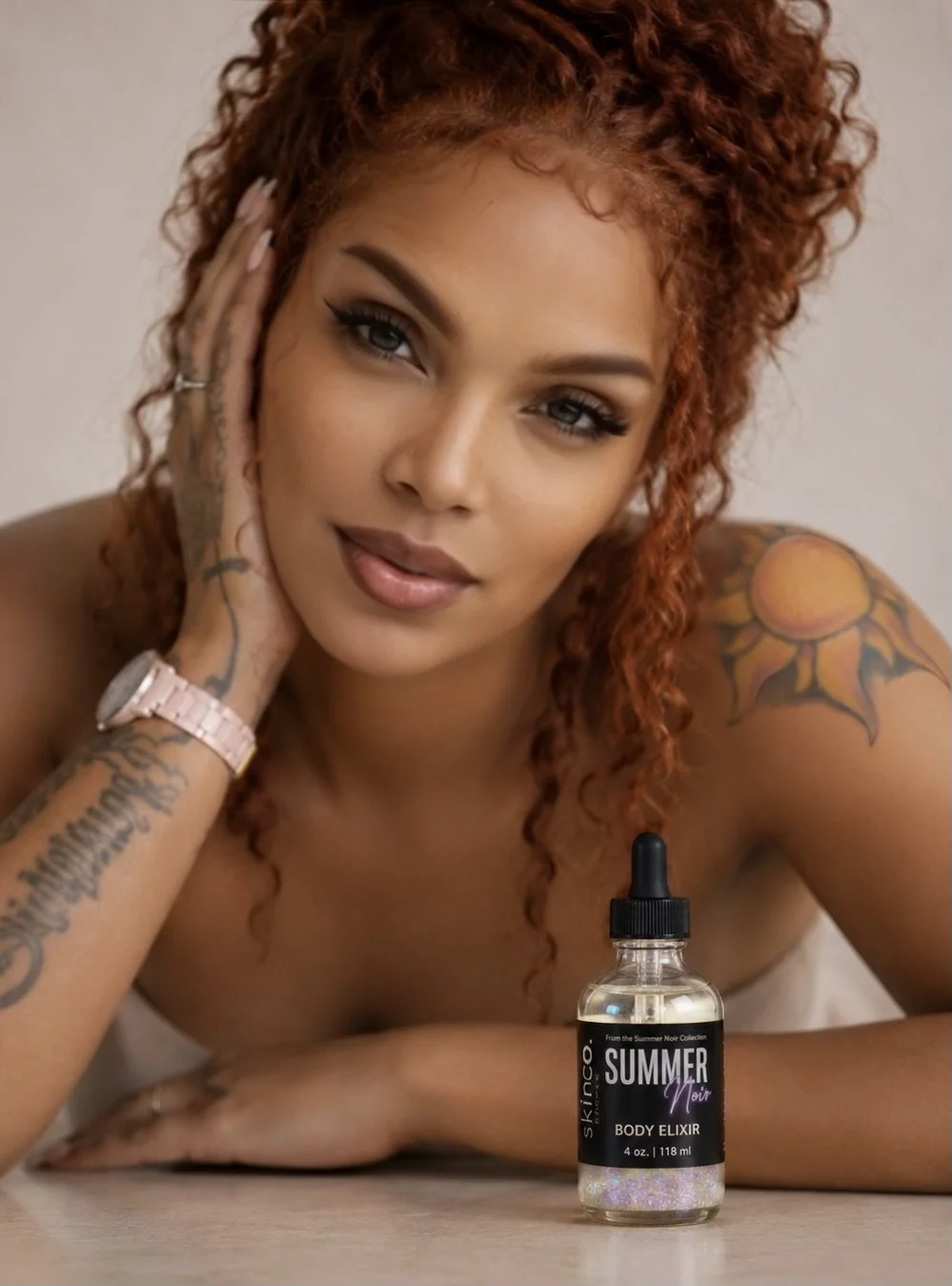 A woman with curly red hair and tattoos, resting her head on her hand, looking at the camera. There is a bottle of Skinn::Summer Mead Body Elixir in front of her on the table.