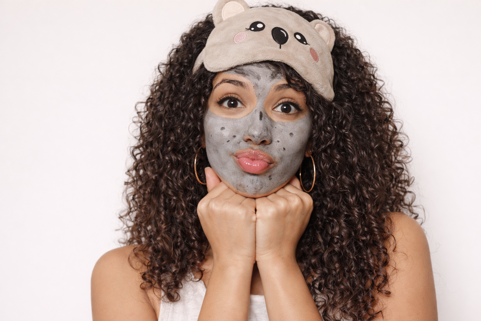 A woman with curly hair wearing a bear-themed sleep mask on her forehead, holding her chin with both hands, and wearing a gray facial mask with dark spots.