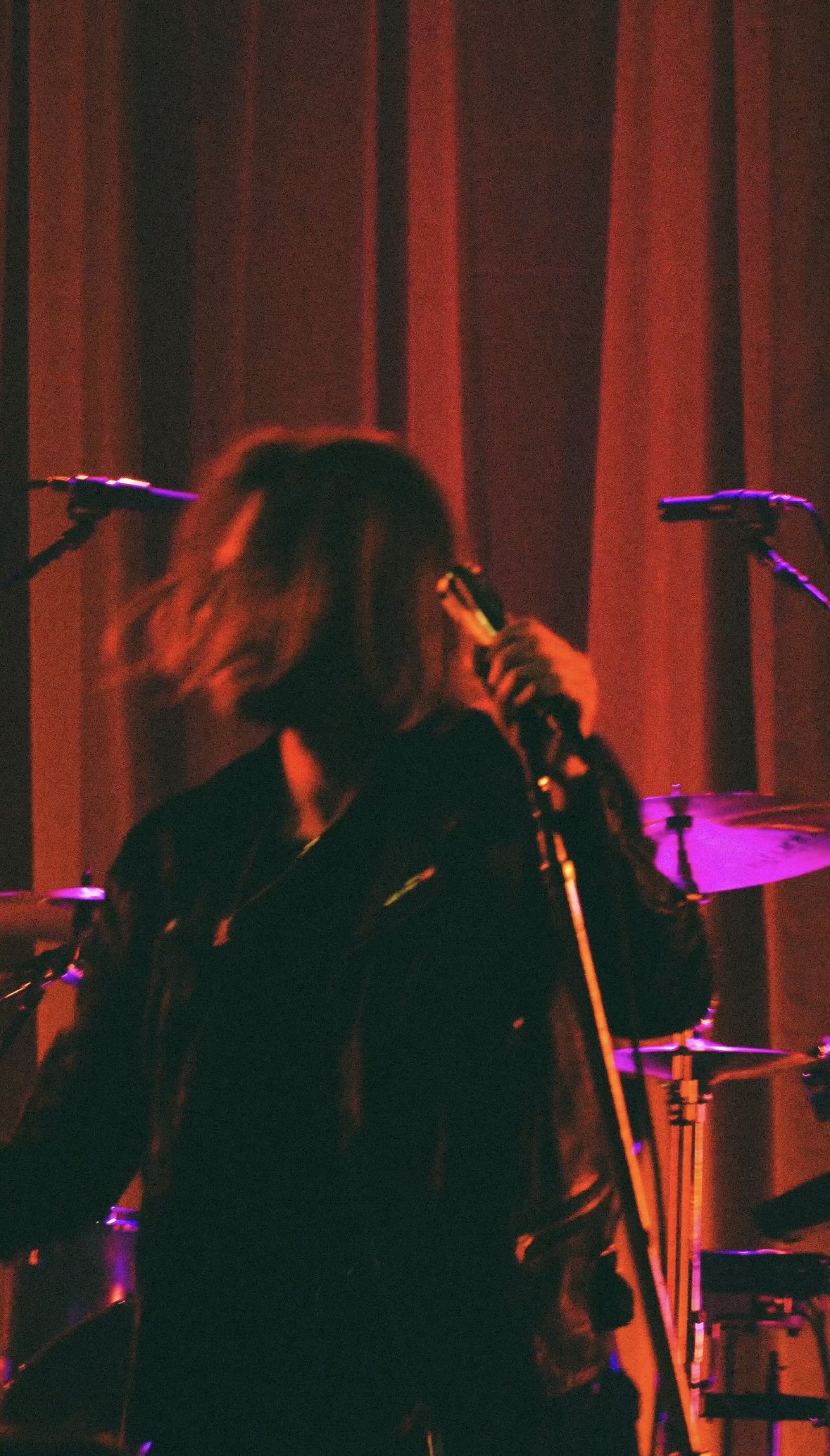 Person with shoulder-length hair holding a microphone on stage with red curtains and musical instruments in the background, illuminated by colorful stage lights.