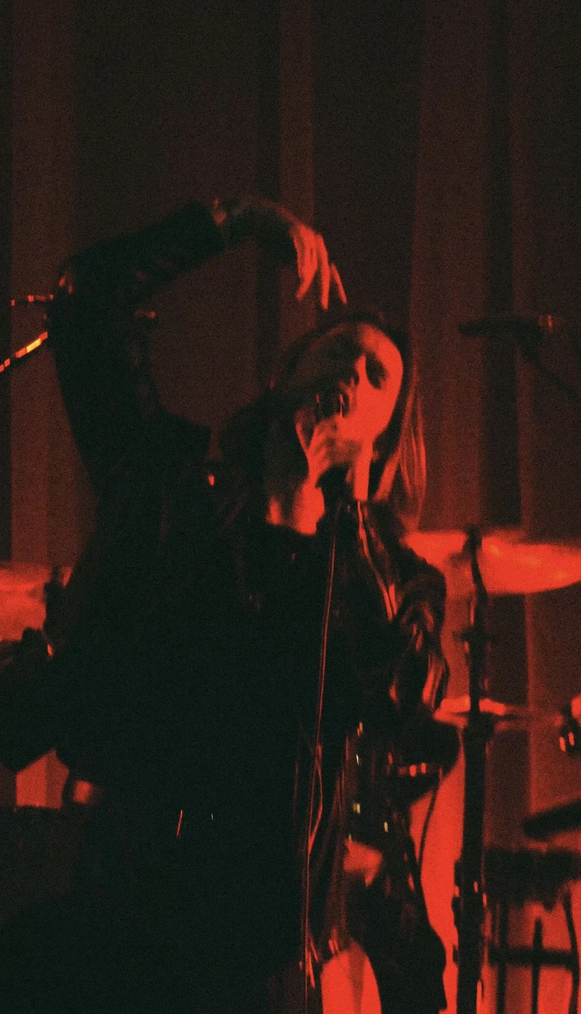 A person performing on stage with red lighting, holding a microphone and gesturing with their hand.