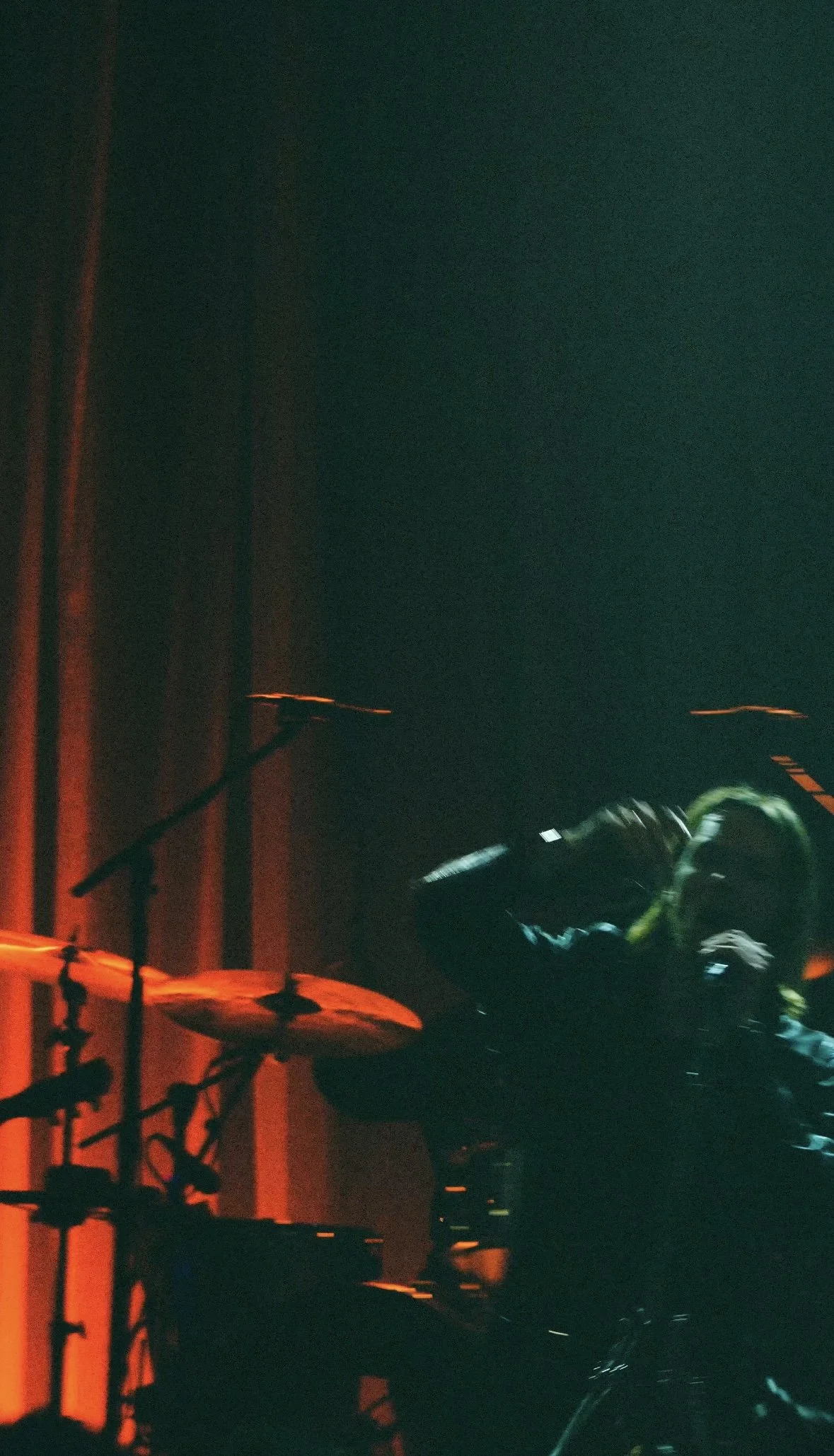 A person wearing headphones and a jacket, sitting at a drum set on a dimly lit stage with red lighting, holding drumsticks.