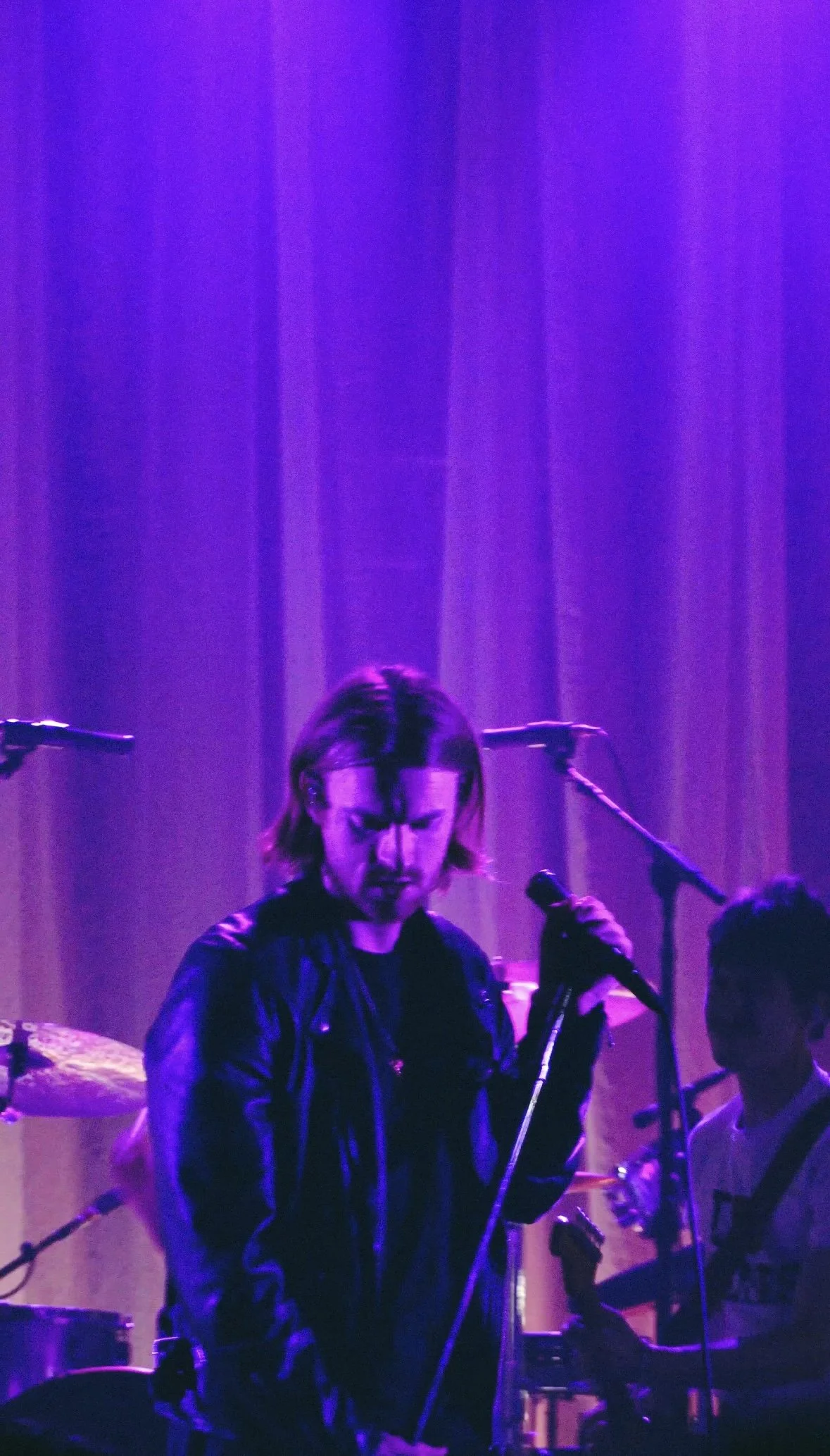 A male musician with shoulder-length hair, wearing a black leather jacket, holding a microphone stand on stage illuminated by purple lighting, with other band members and instruments in the background.