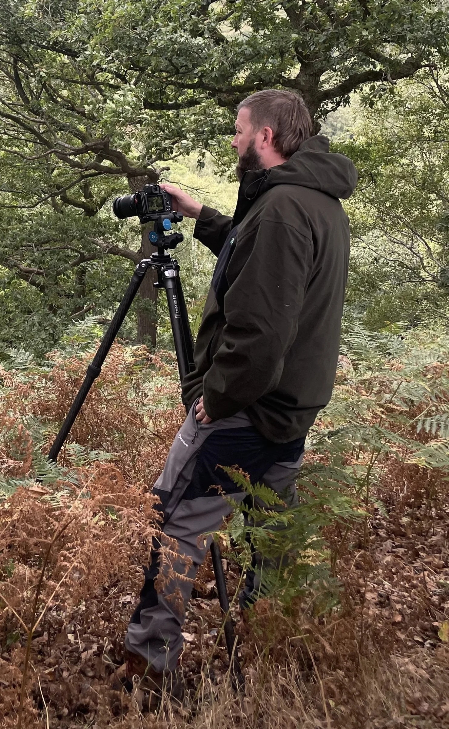 A man in outdoor clothing stands in a forested area, adjusting a camera mounted on a tripod.