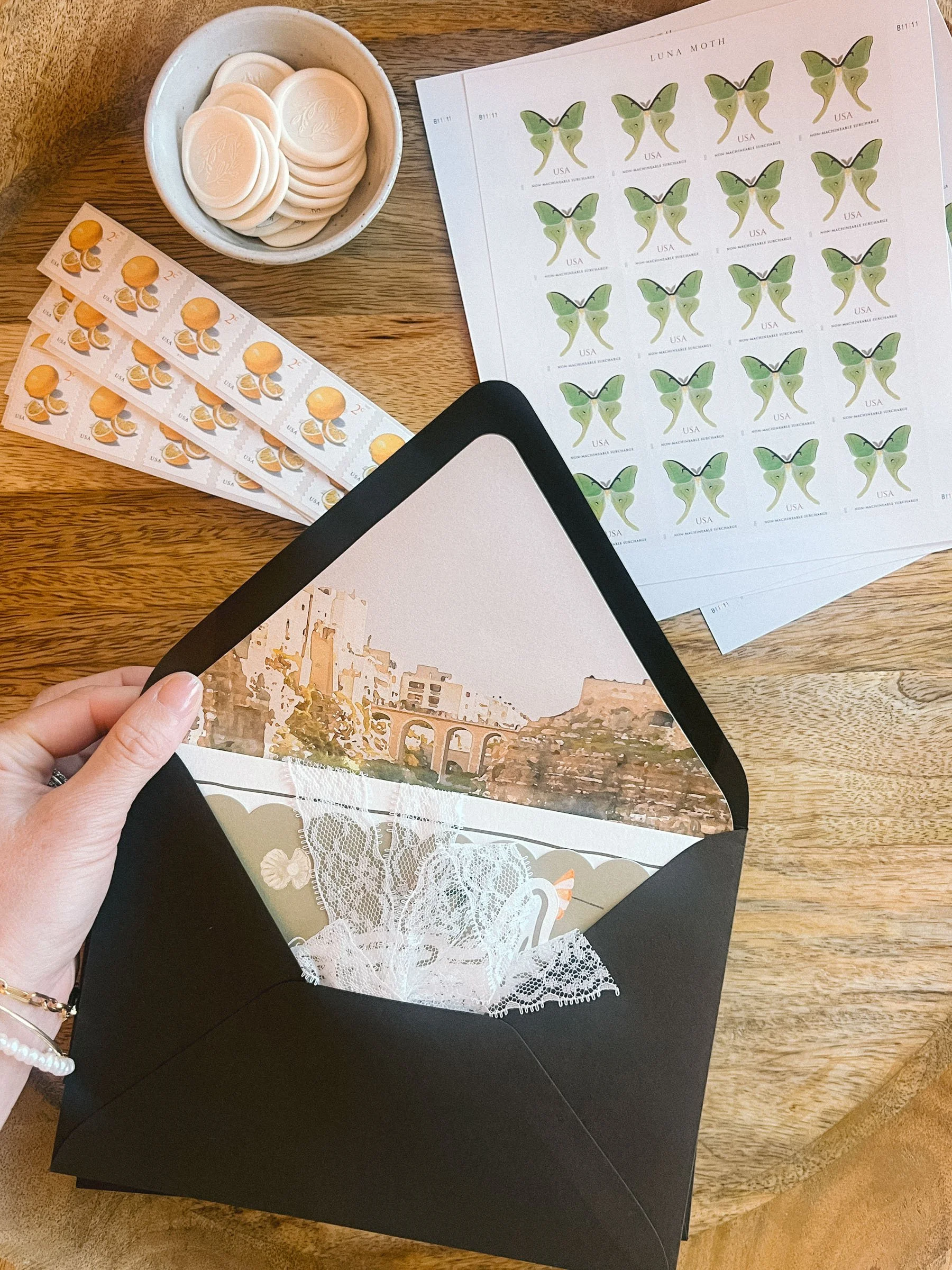 A person's hand holding a black envelope with lace and a picture of a cityscape inside. On the table are sheets of stamps featuring a butterfly and a lemon dessert, a dish with white circular objects, and a sheet of butterfly stamps.