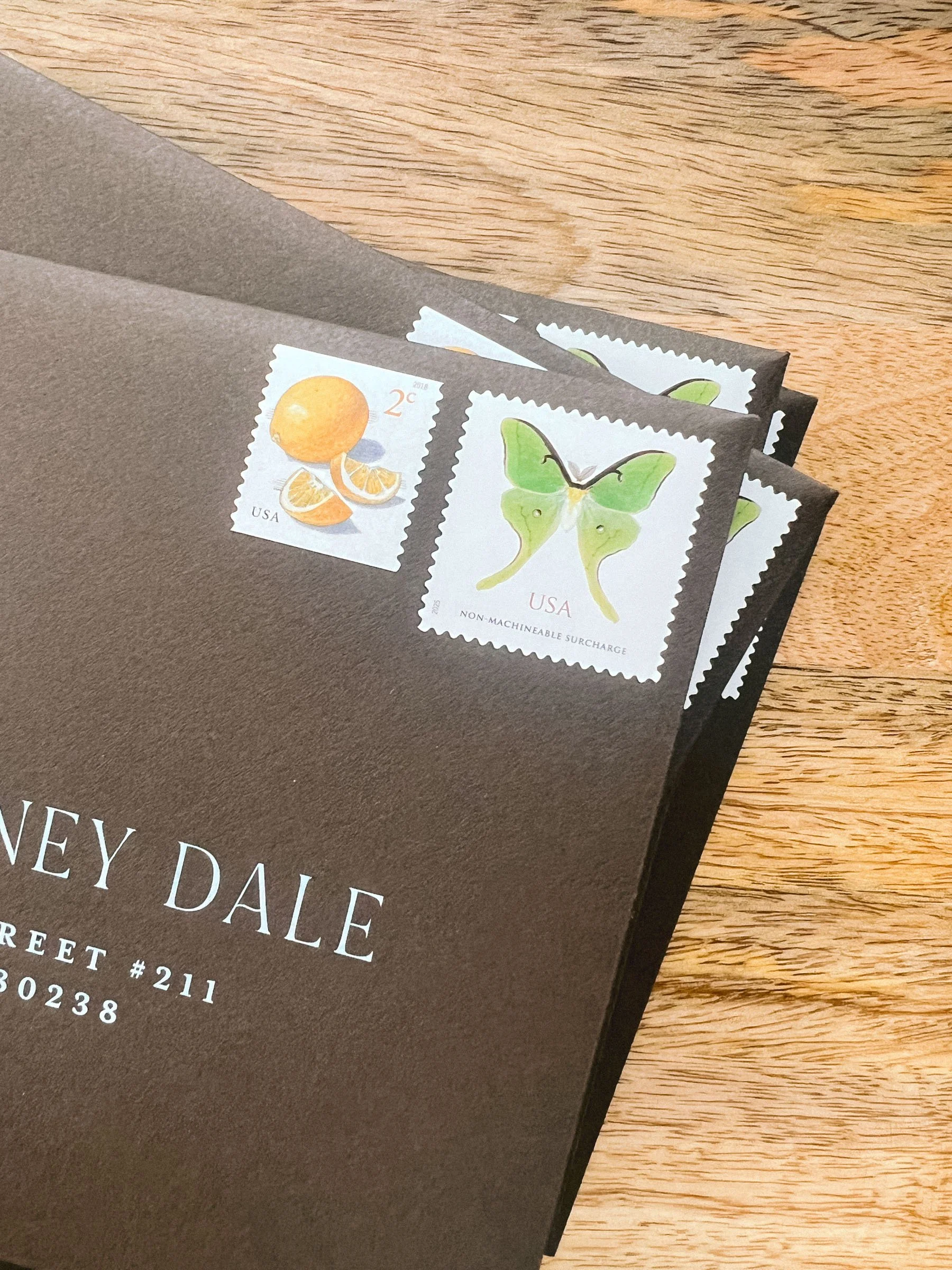 A dark brown envelope with three USA postage stamps featuring a lemon and a butterfly, lying on a wooden surface.