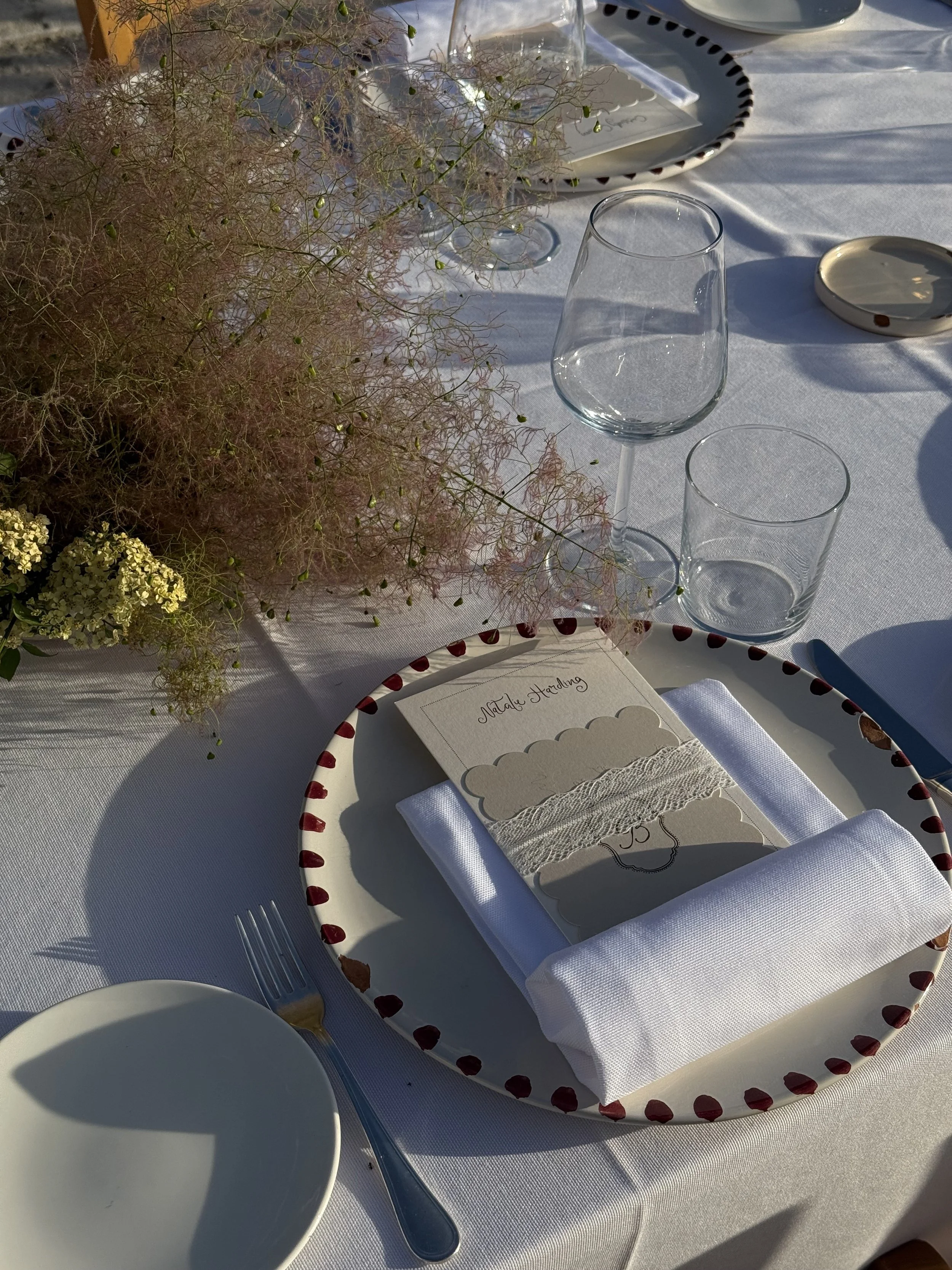 Elegant table setting with a white dinner plate, white napkin, utensils, and a menu card, with a flower arrangement and glassware in the background.