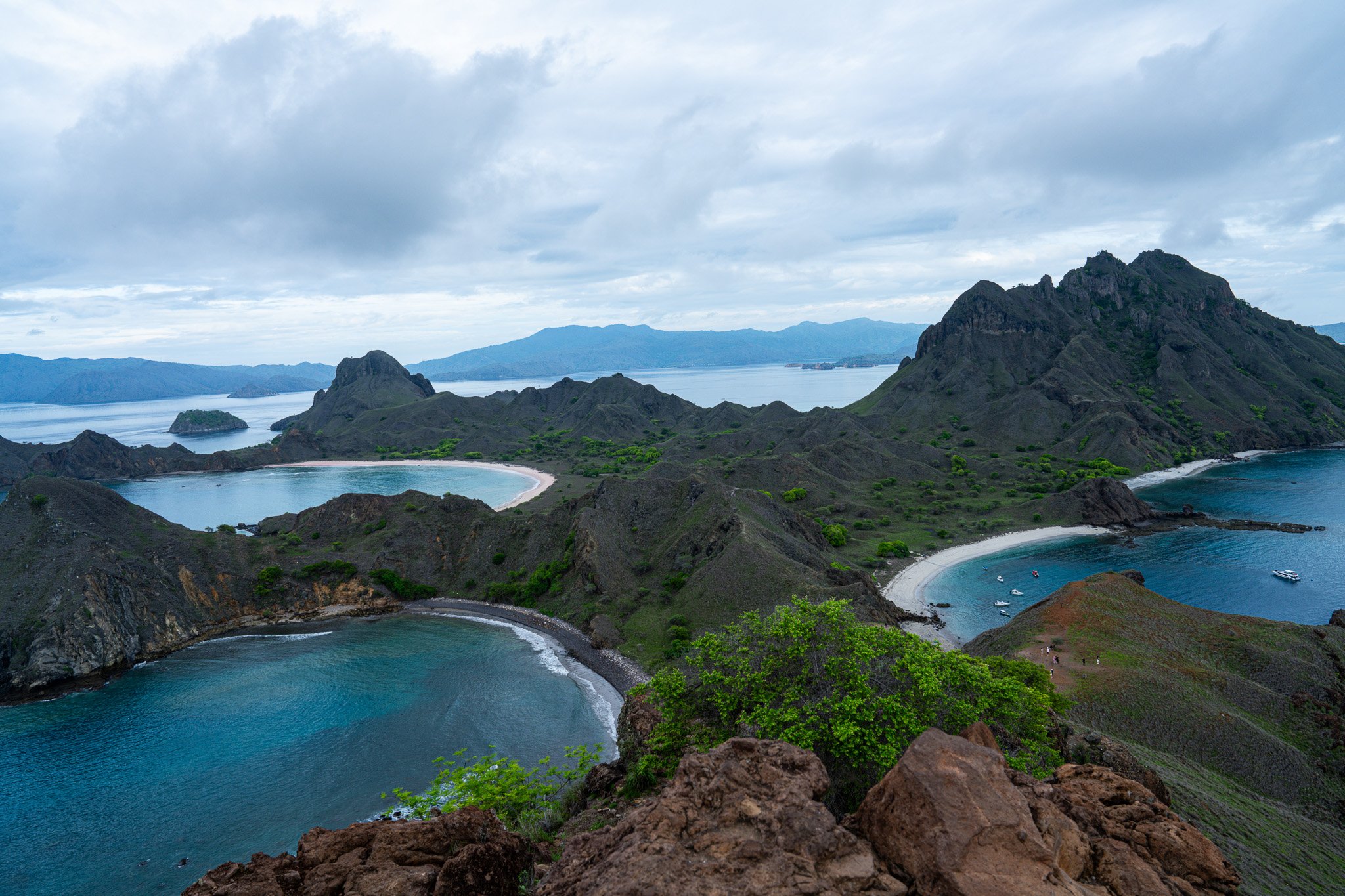 Scenic view of a landscape with multiple small bays and rugged green hills under a cloudy sky, with boats anchored in the water.