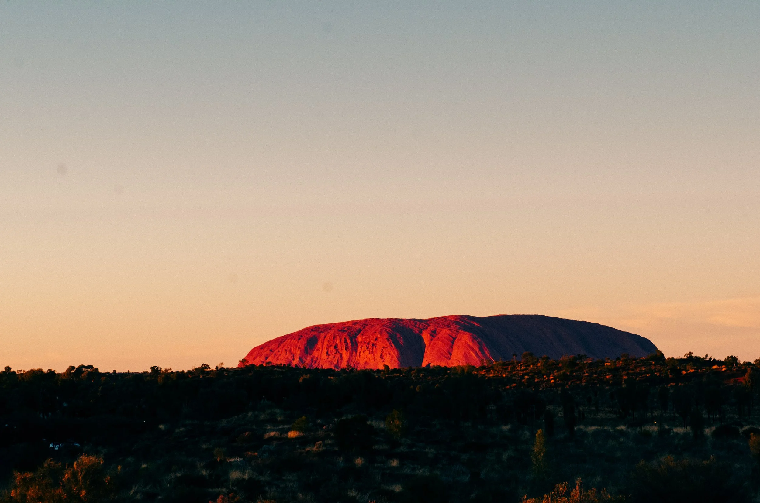 Sunset-lit rock formation in a vast landscape with sparse trees and bushes in the foreground.