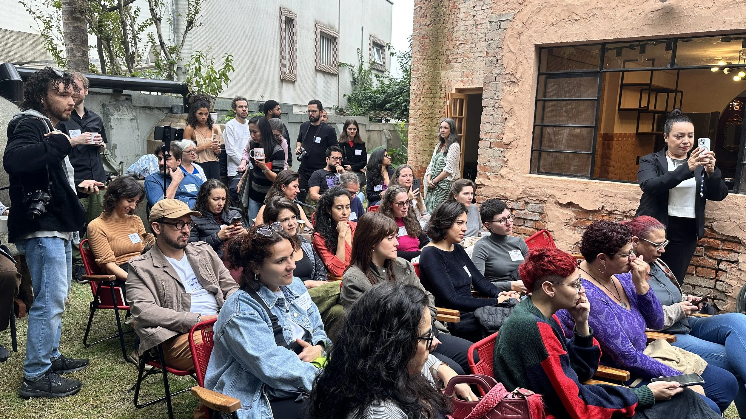 Público de mulheres e homens assistindo a uma apresentação ao ar livre, com algumas pessoas usando camisetas com nomes e sentadas em cadeiras vermelhas, ao lado de uma parede de tijolos expostos, enquanto outras estão de pé com câmeras e celulares registrando o momento.
