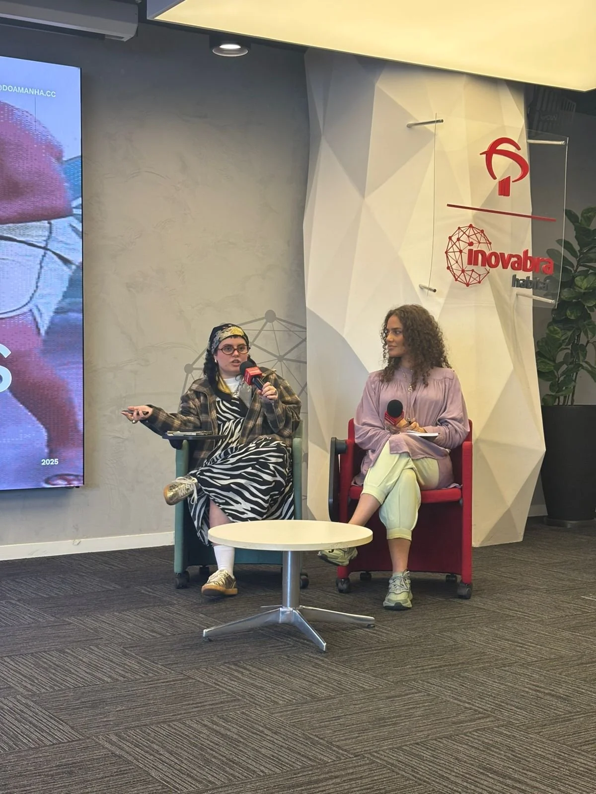Duas mulheres sentadas em cadeiras vermelhas, uma delas fala ao microfone enquanto a outra ouve, em um ambiente de escritório com parede decorada, logo da Inovabra Habitat e uma tela ao lado