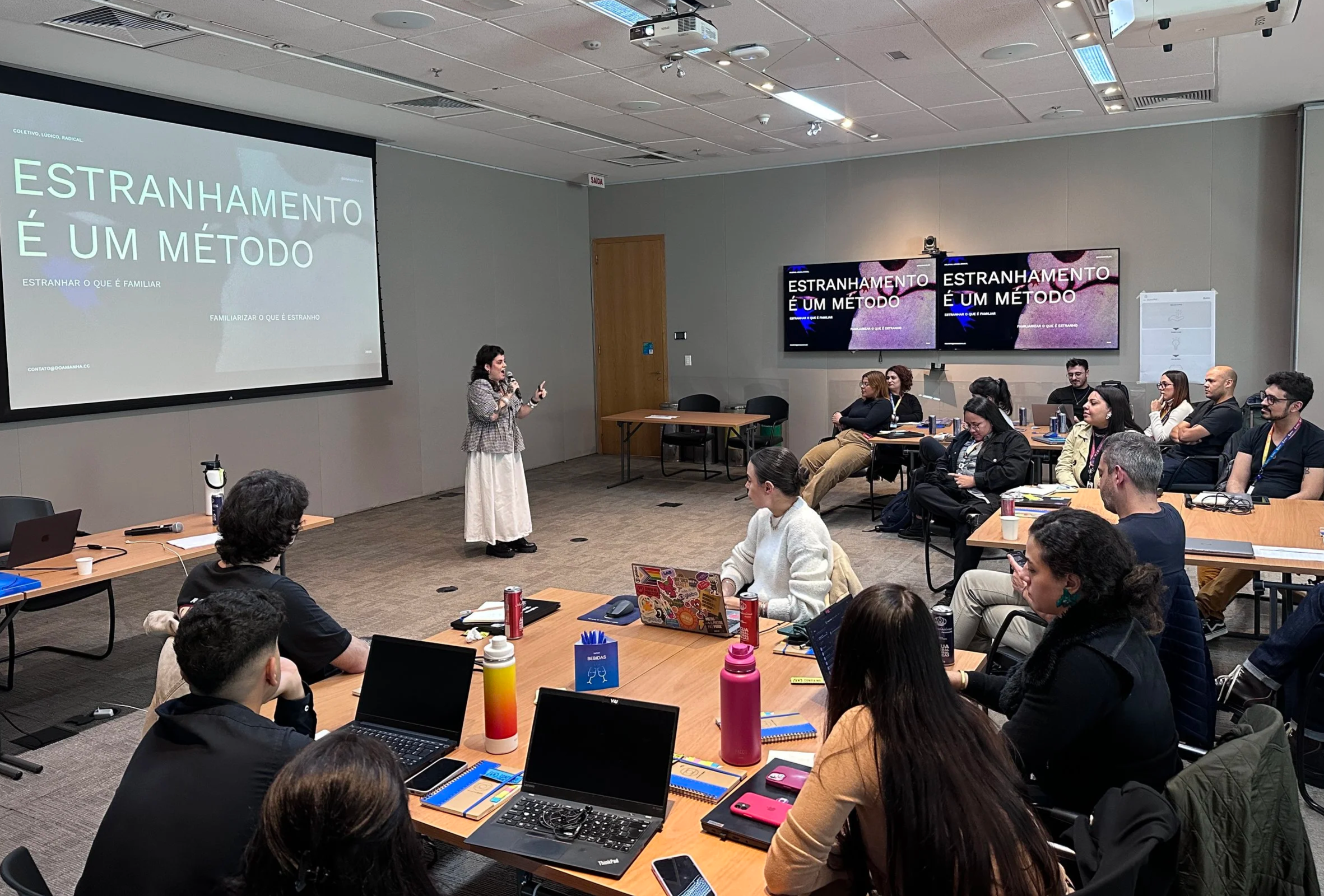 Pessoa apresentando em uma sala de aula ou conferência, com uma projeção na parede que diz 'Estranhamento é um método', enquanto os participantes assistem e utilizam computadores e materiais de escritório.