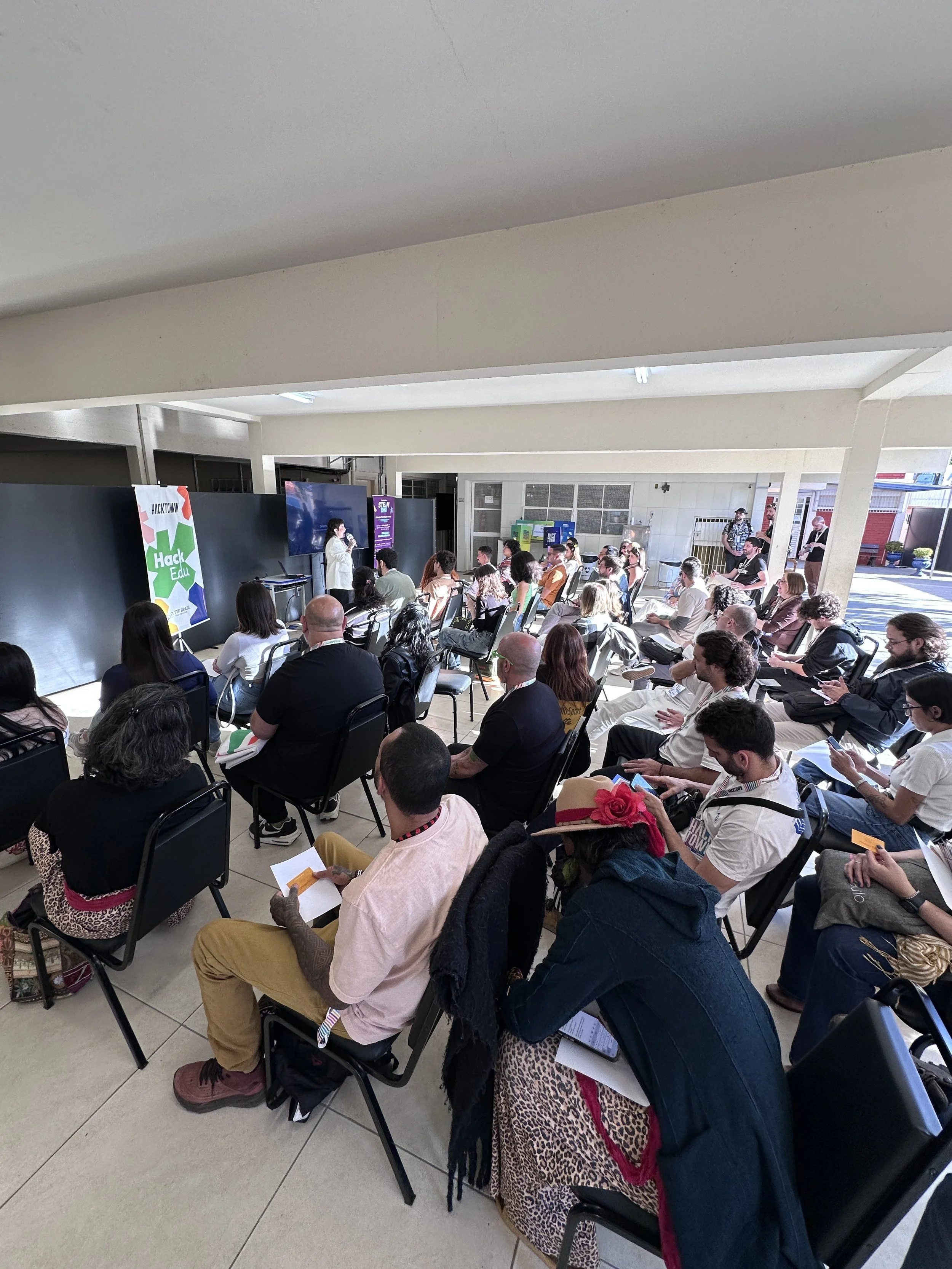 Palestra com uma palestrante falando em um palco com máscara na face, enquanto a audiência assiste, sentada em cadeiras, em uma sala bem iluminada ao ar livre.