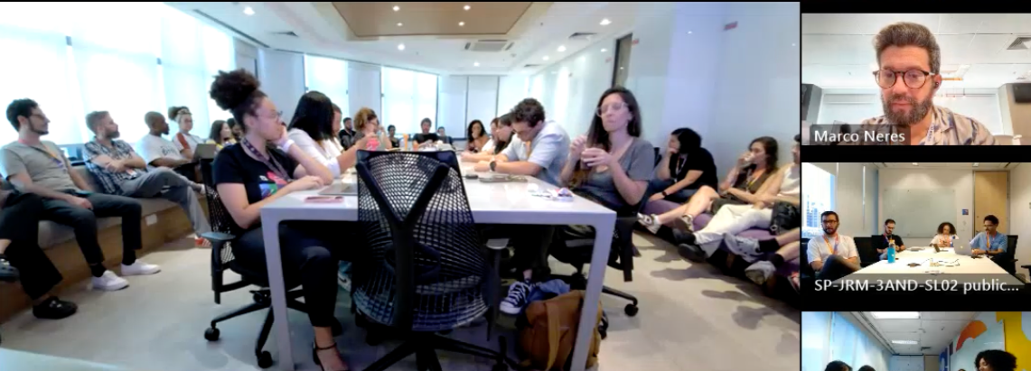 Reunião ou workshop com várias pessoas sentadas ao redor de uma mesa em uma sala bem iluminada, com janelas grandes. Alguns participantes estão usando laptops e roupas casuais, enquanto uma pessoa está falando e segurando um copo. A tela mostra uma videochamada com duas outras pessoas incluindo uma identificada como Marco Neres.