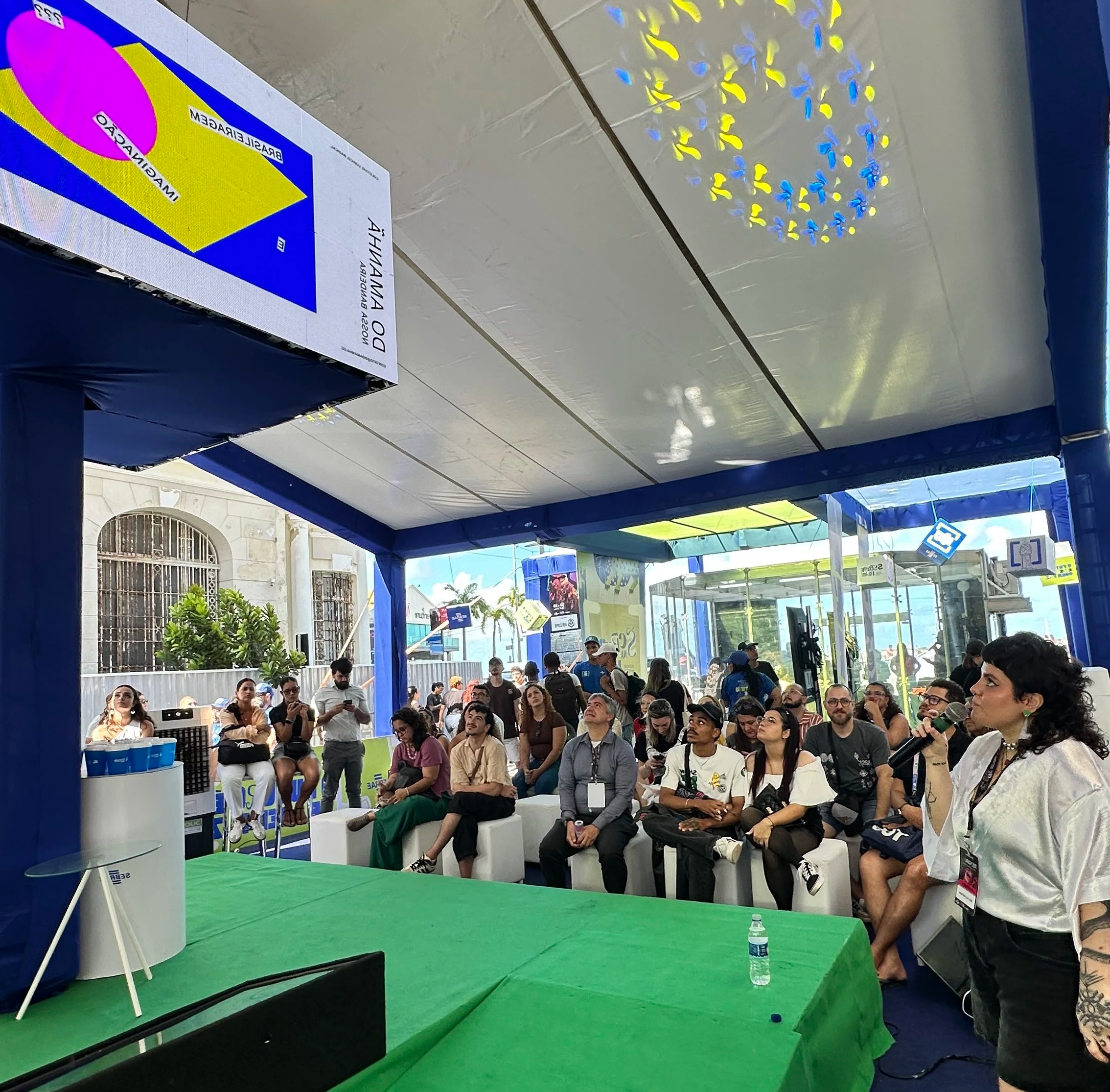 Pessoas sentadas em uma sala com toldos azuis, participando de evento com uma mesa coberta por uma toalha verde, algumas pessoas observando uma mulher com microfone.