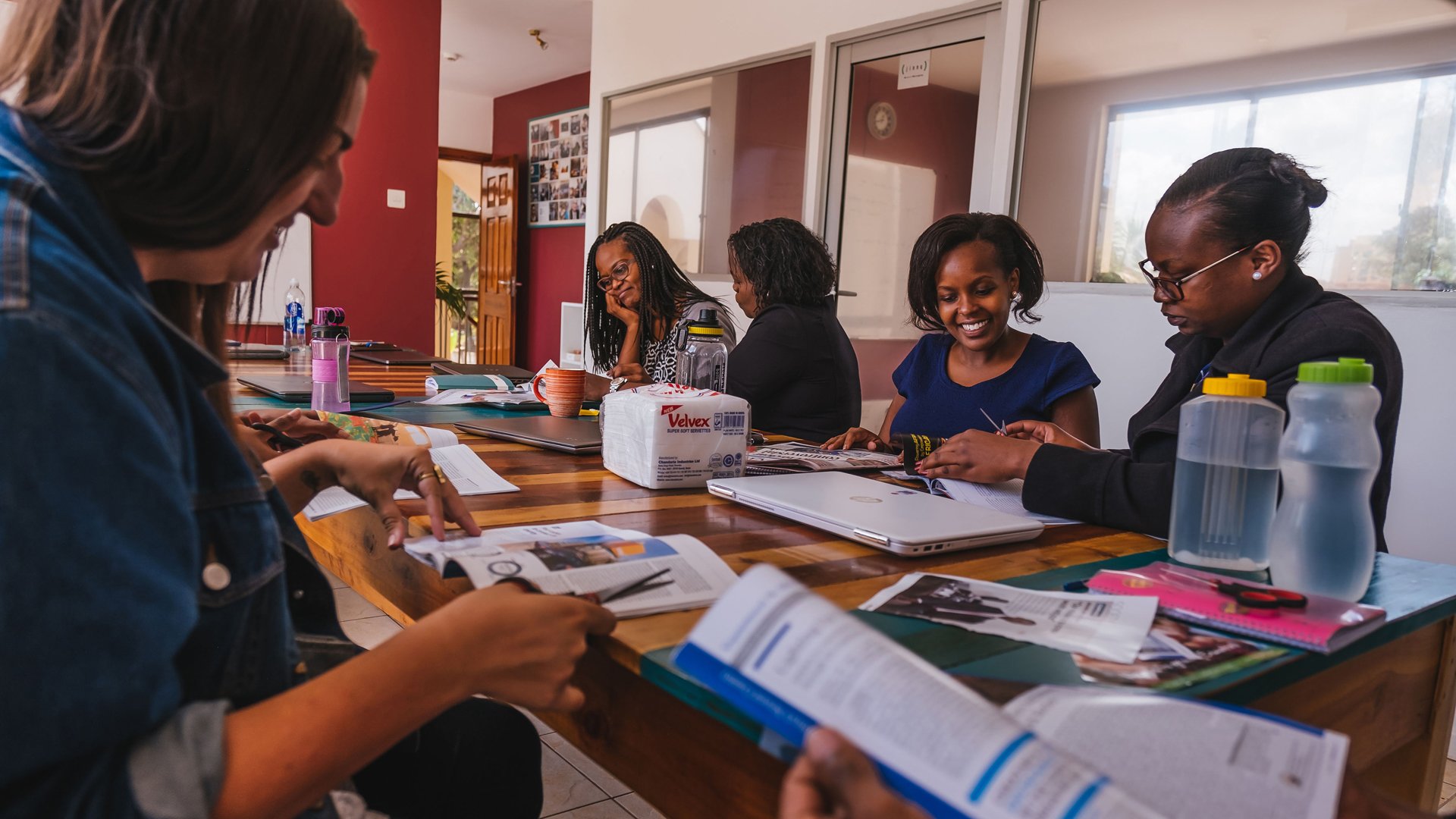 Group of women sitting around a wooden table in a meeting room, reading magazines and documents, with laptops, water bottles, and a pack of Velvex tissue on the table, and a wall with photos and a window in the background.