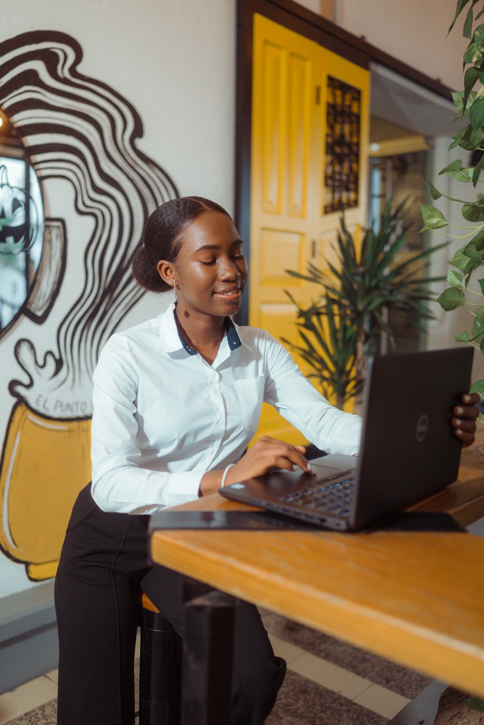 A woman with dark hair styled in a bun, wearing a white blouse and black skirt, working on a laptop at a wooden counter in a café. Behind her, there is a yellow door and wall art featuring a black and white illustration of a face with flowing lines. Potted plants are also visible.