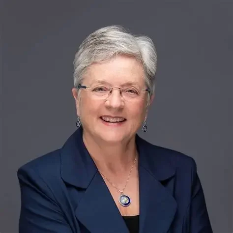 Smiling elderly woman with short gray hair, glasses, wearing a navy blazer and jewelry, against a gray background.