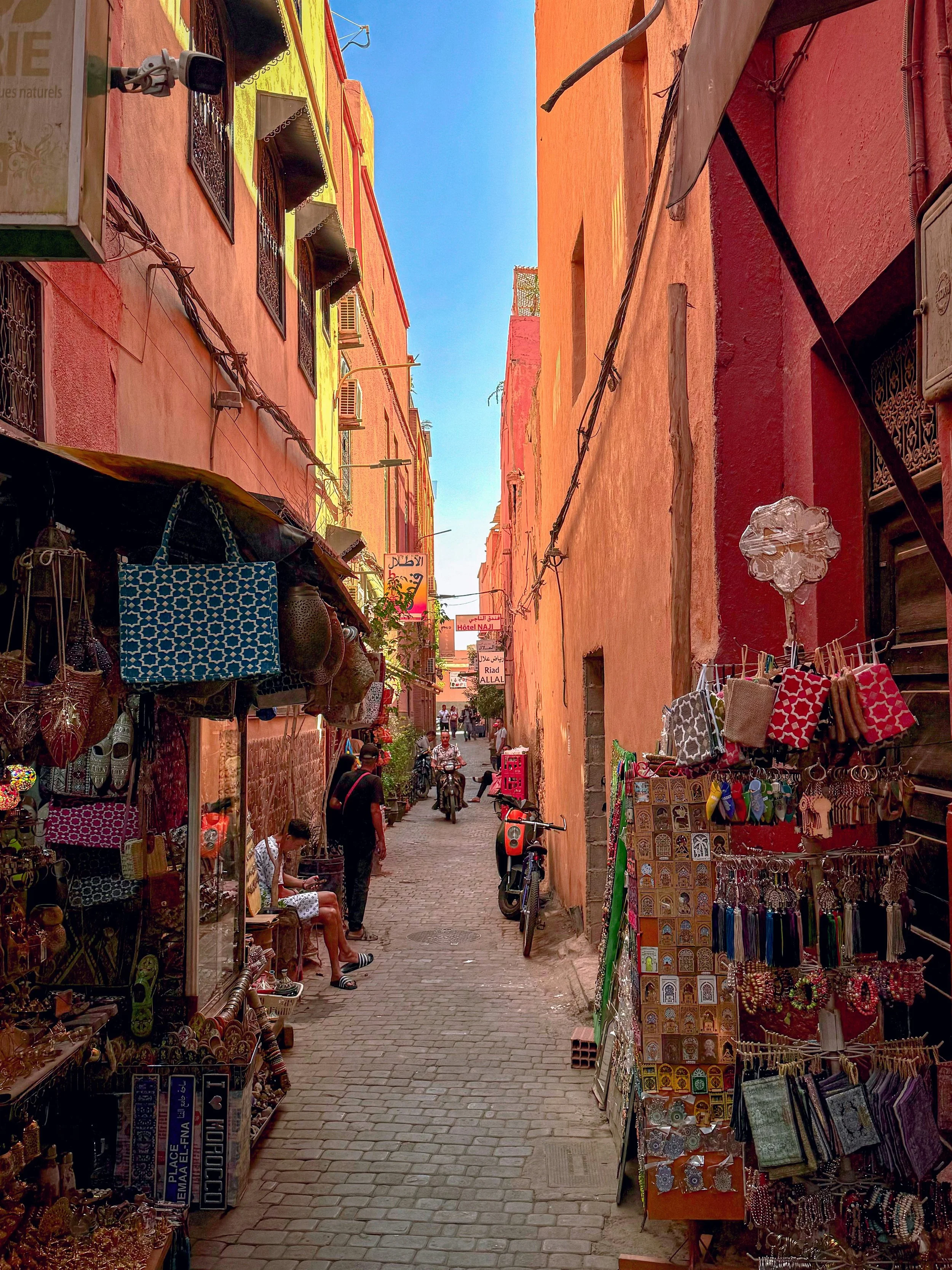 A narrow alleyway in a market with colorful buildings on both sides, shop displays with jewelry, bags, and souvenirs, and some pedestrians and bicycles in the street.