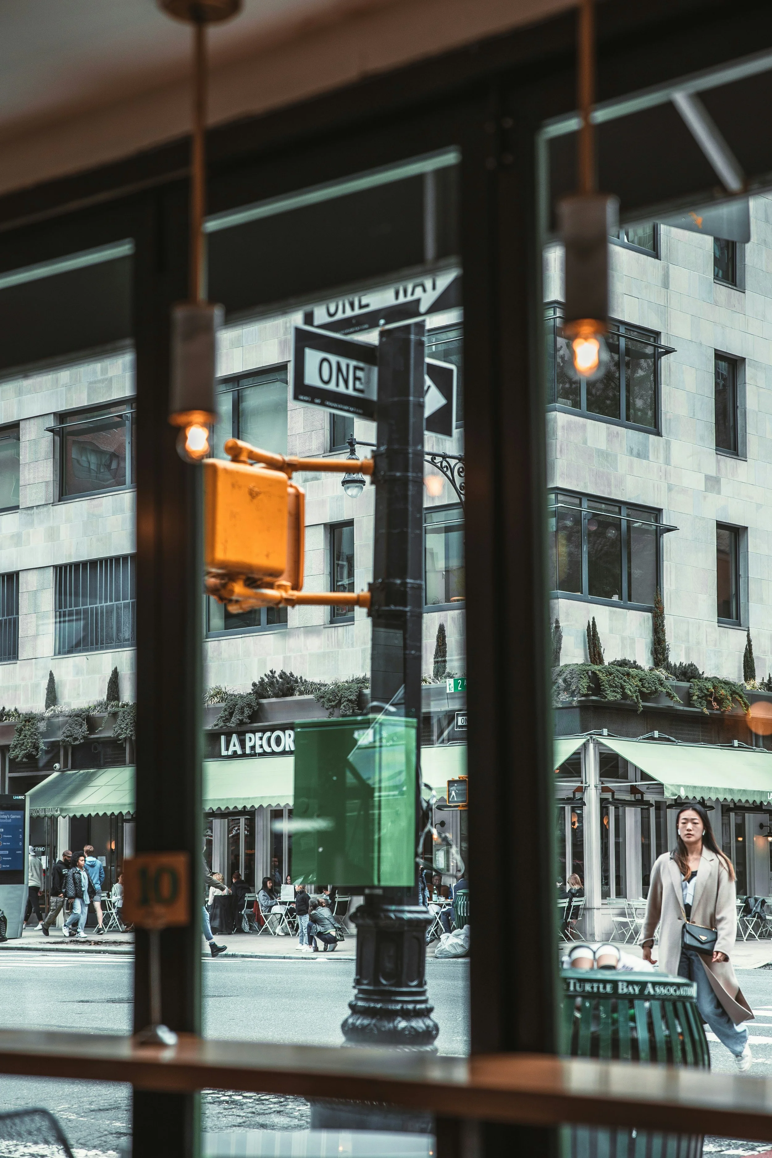 Urban street scene viewed through a window with hanging lights. People walk along the sidewalk in front of a modern building with large windows and outdoor seating. A woman wearing a beige coat walks past a green bench labeled 'Turtle Bay Association.' Street signs indicate a one-way street, and a yellow traffic signal is visible.