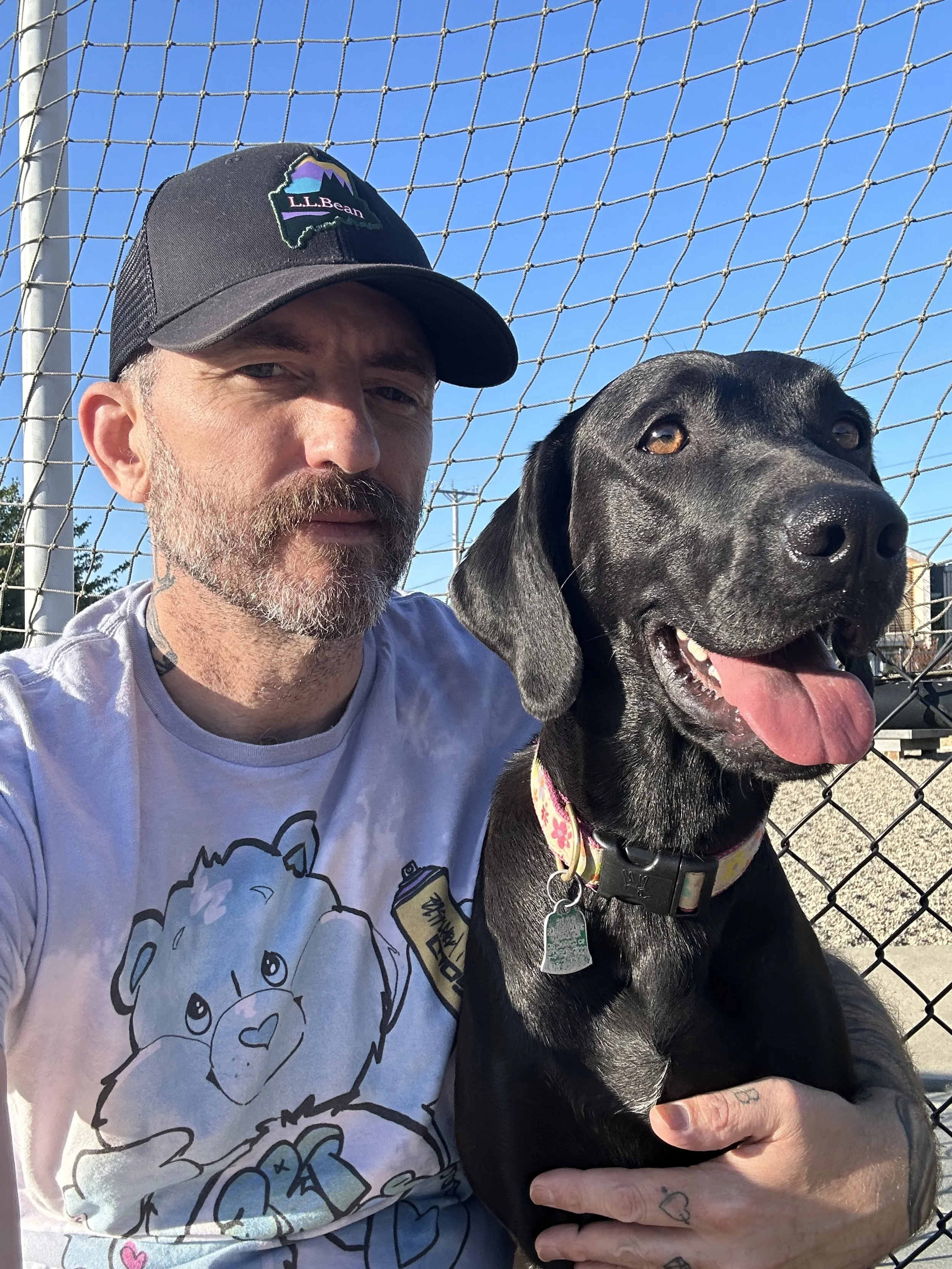 A man with a beard and a tattoo on his hand, wearing a baseball cap and a tie-dye T-shirt, taking a selfie with a happy black Labrador Retriever dog in front of a chain-link fence on a sunny day.