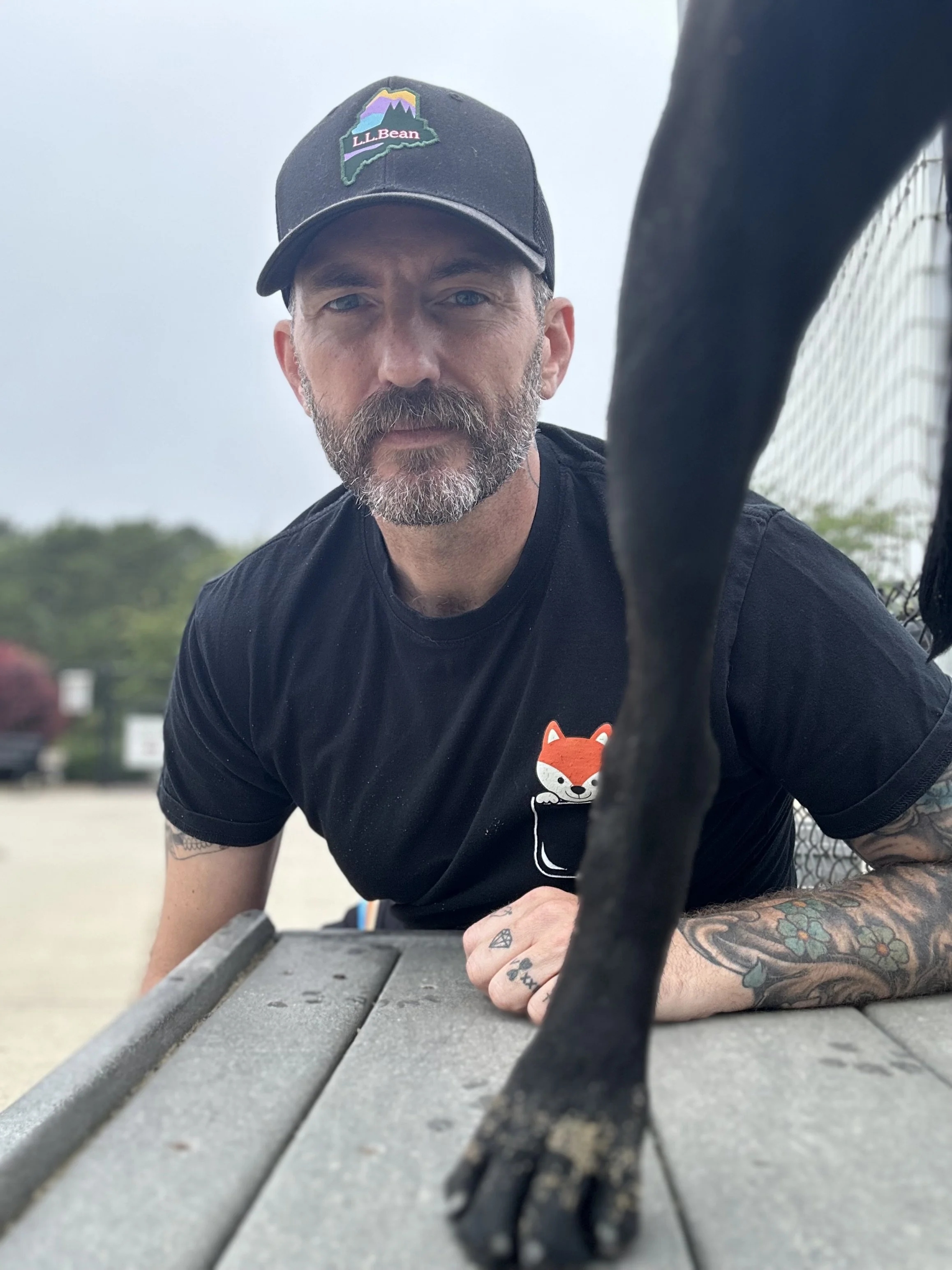 A man with a beard and tattoos wearing a black T-shirt with a fox graphic and a black cap, leaning over a picnic table with a black dog, outdoors during daytime.