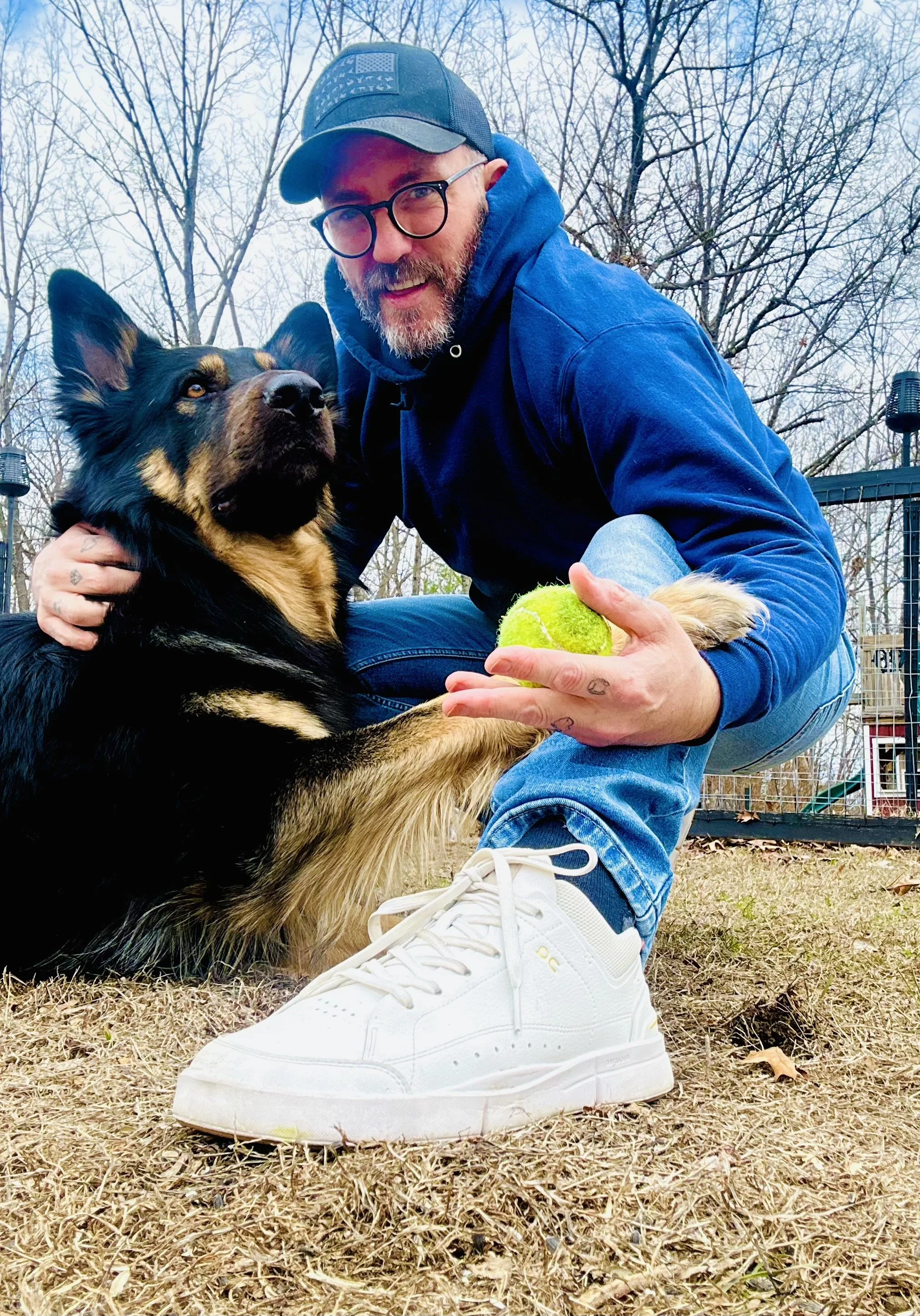 A man wearing glasses, a baseball cap, blue hoodie, and jeans crouches on the ground outdoors with a large dog, holding a tennis ball, in a fenced yard with trees in the background.