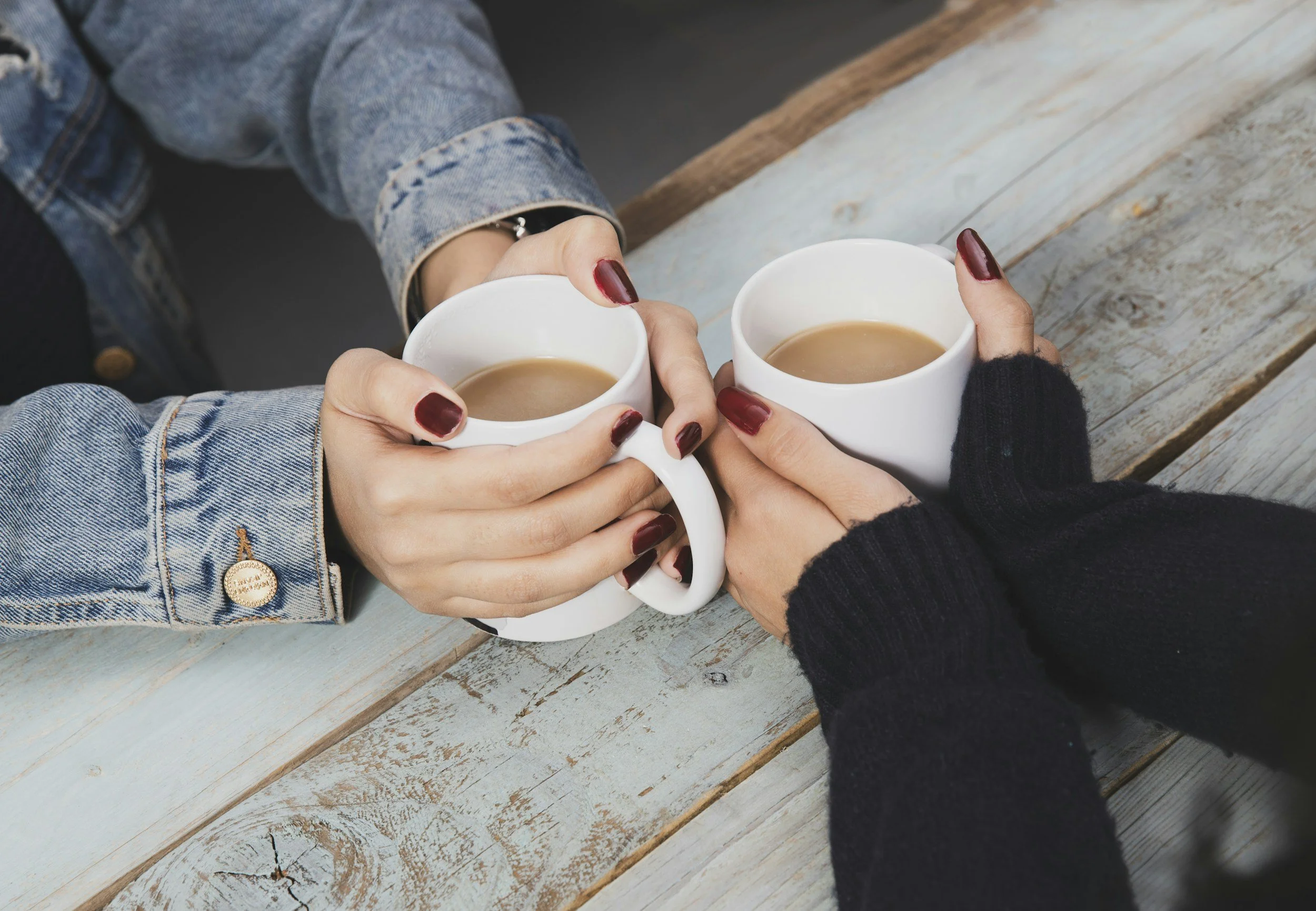 Two people holding white mugs of coffee, clinking them together on a rustic wooden table. One person wears a denim jacket with a gold button, and the other wears a black sweater.