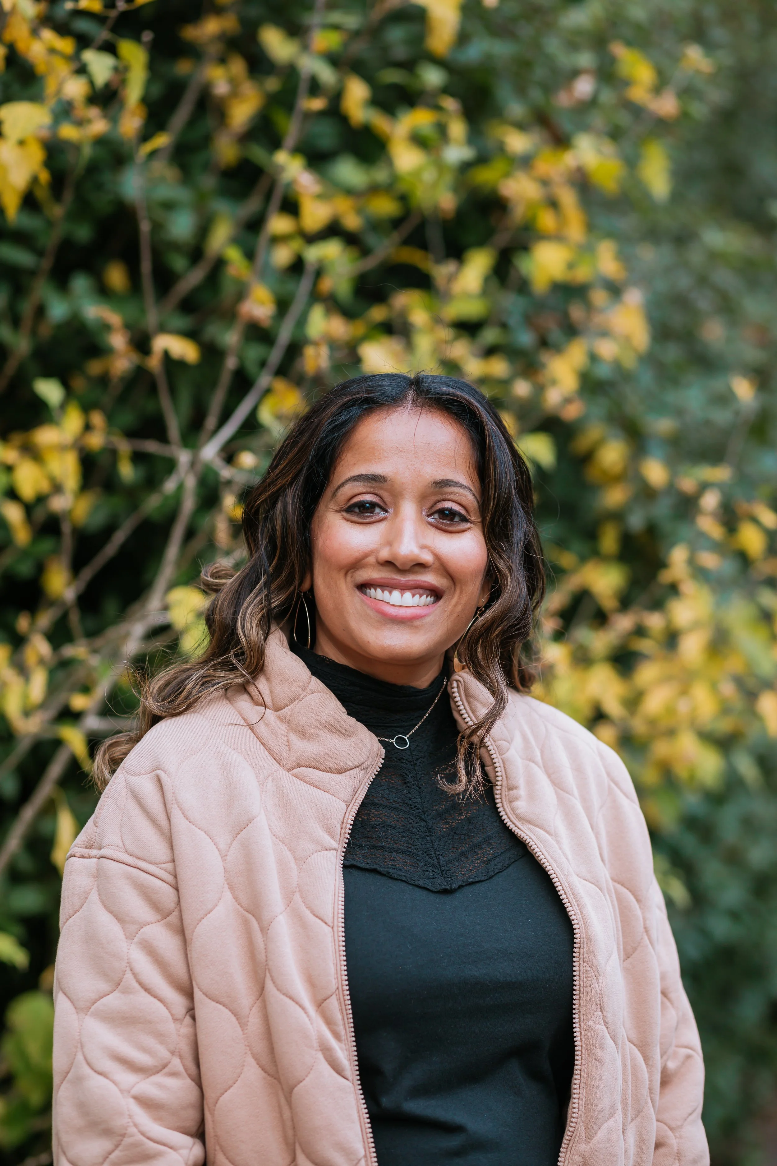 A woman smiling outdoors wearing a beige quilted jacket and black top, with a background of autumn yellow and green leaves.
