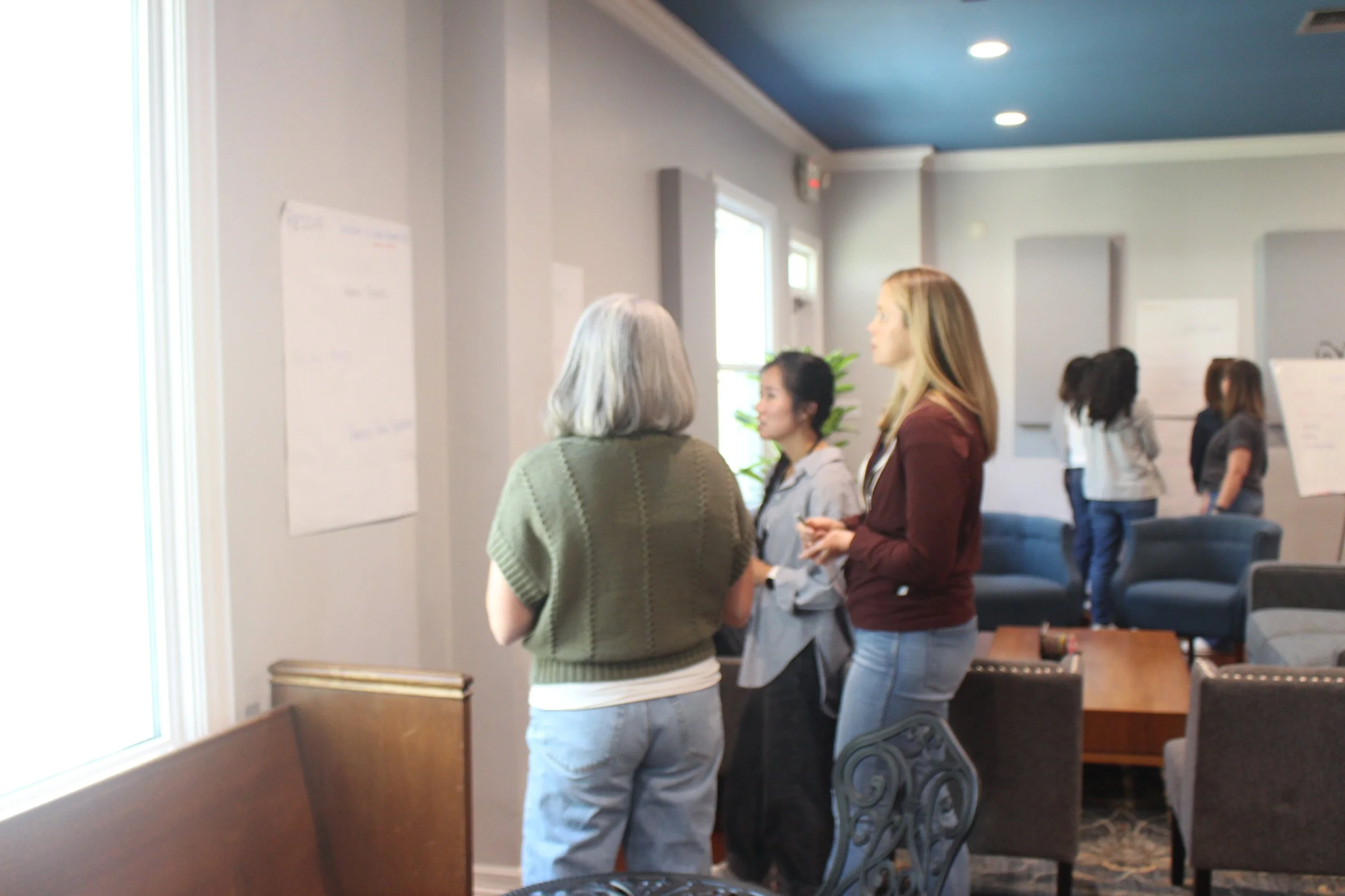 A group of women are standing and talking in a conference or meeting room with gray walls and a blue ceiling. There are chairs and whiteboards in the background.