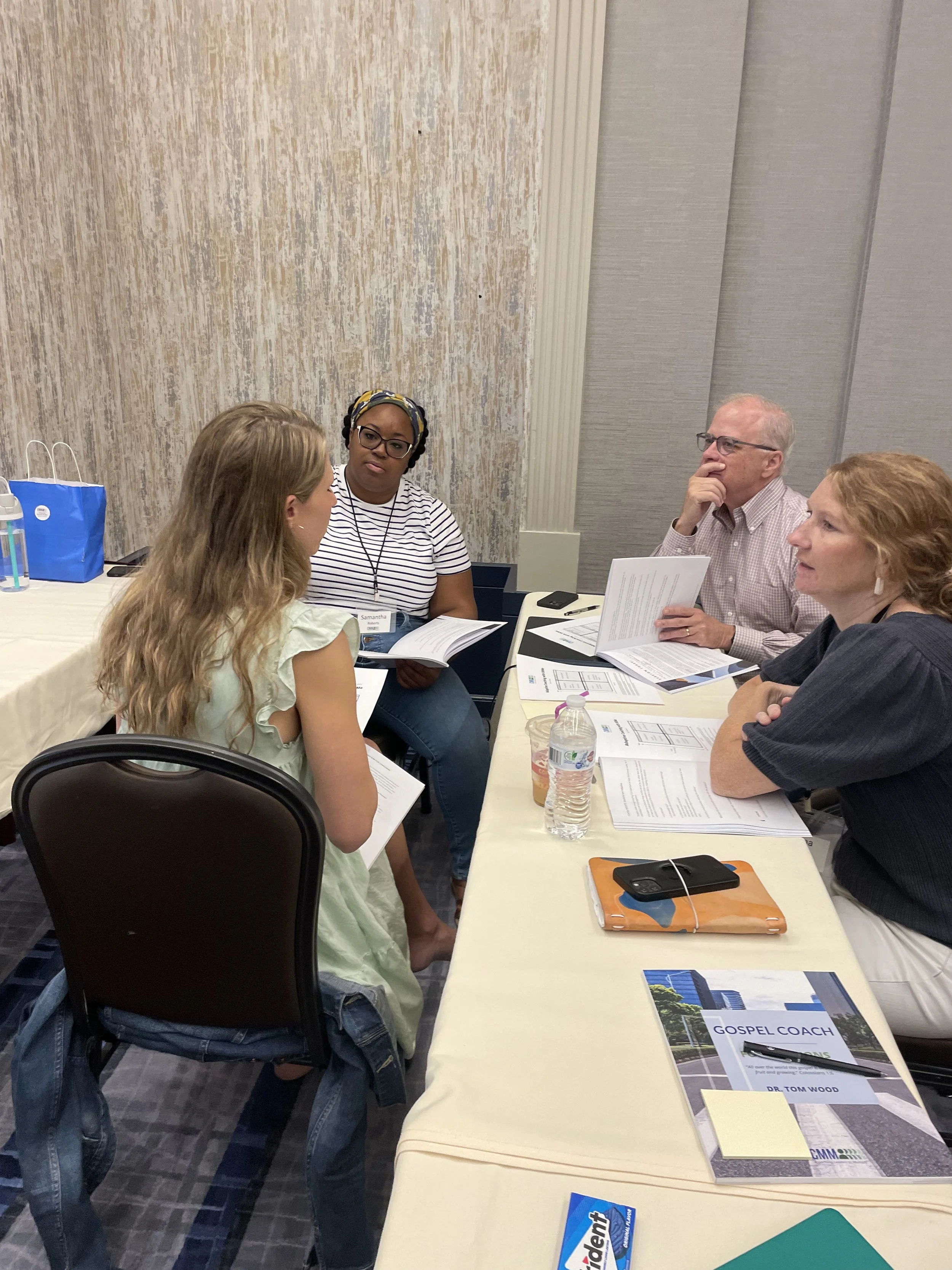 Four people are sitting around a table engaging in a discussion at a Gospel Coaching Foundations Training, with papers, a water bottle, and a tablet on the table.