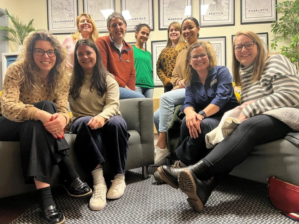 Group of nine women sitting and standing in a room with framed city maps on the wall and potted plants, smiling and posing for the photo.