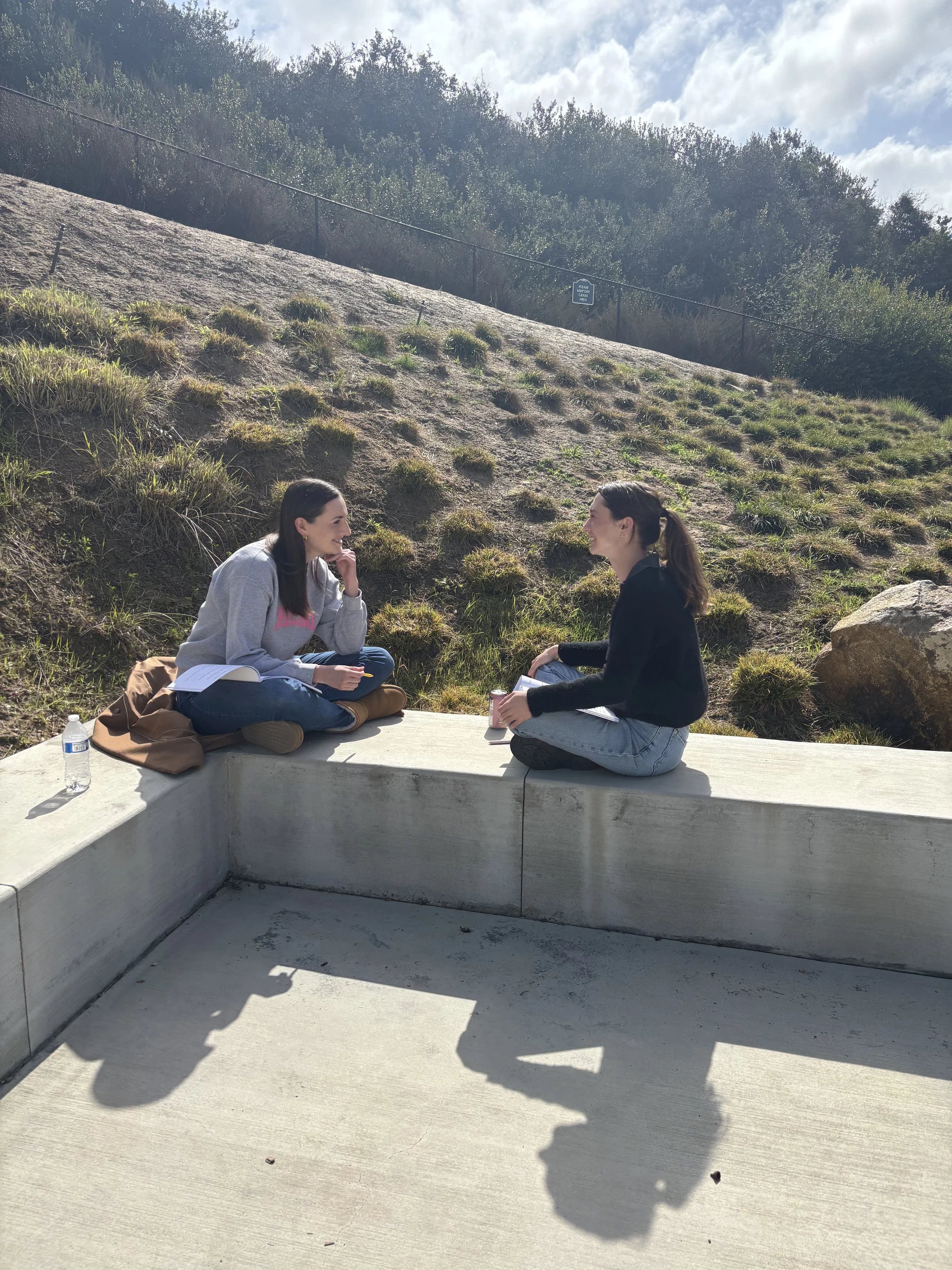 Two women coaching one another on a concrete ledge outdoors, engaged in conversation, with notebooks and water bottles nearby, on a hillside with grass and bushes under a partly cloudy sky.