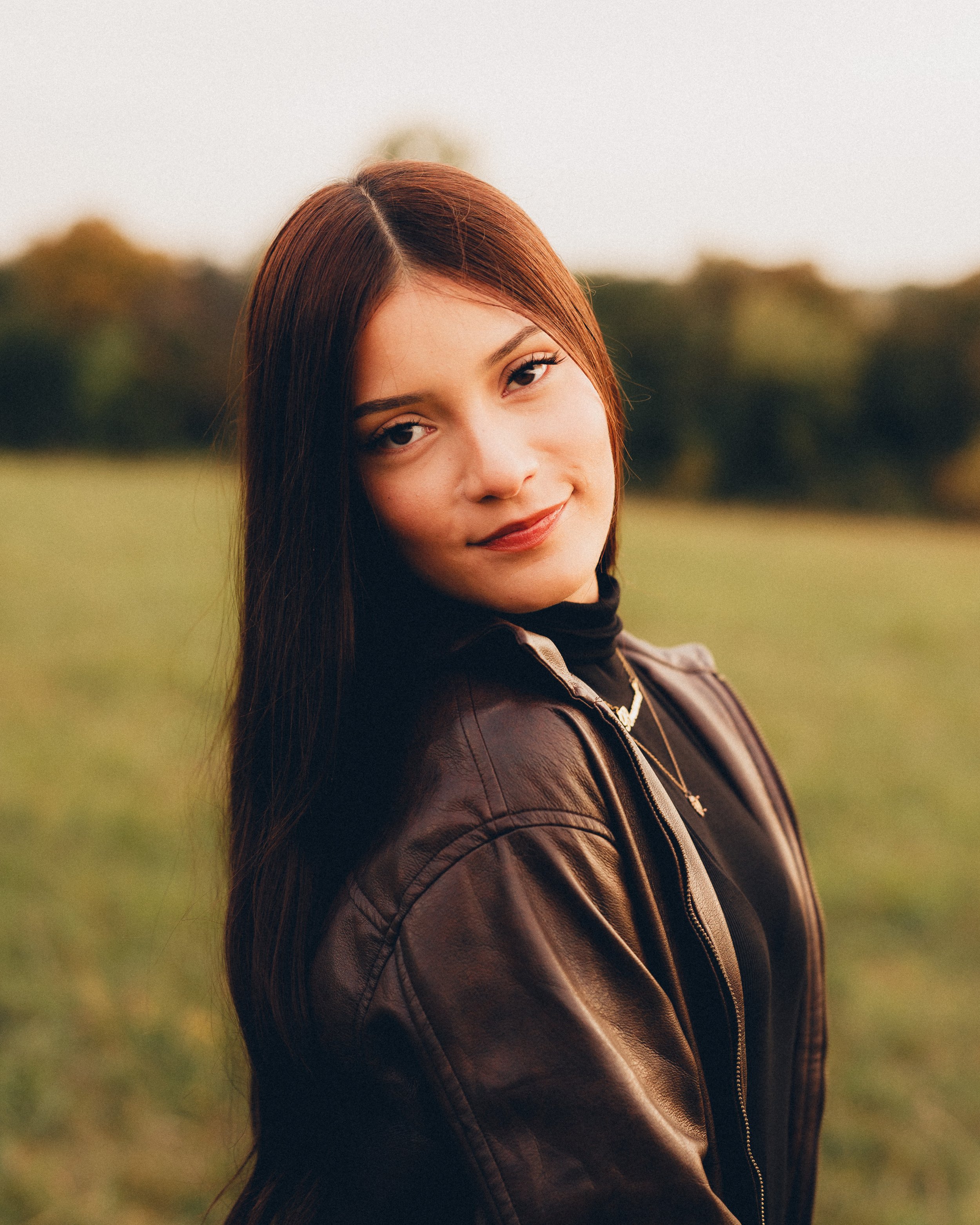 A young woman with long brown hair, wearing a black turtleneck and a brown leather jacket, outdoors in a grassy field during golden hour, looking at the camera with a slight smile.