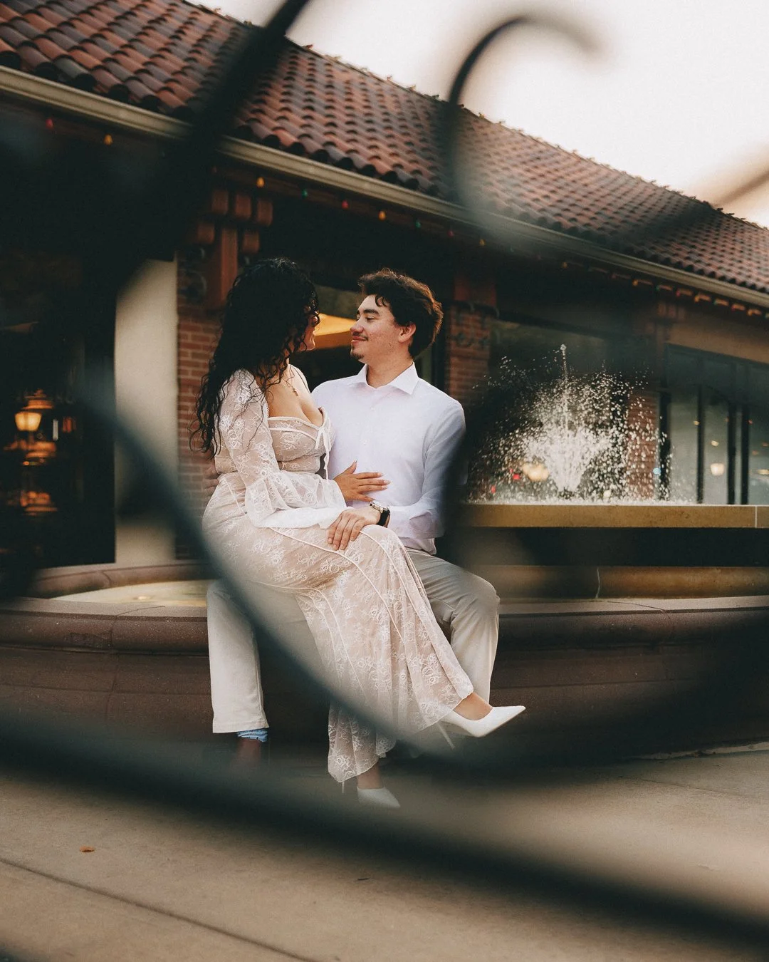 A couple sitting on a bench at a fountain outside a brick building during sunset, looking at each other affectionately. Shot at Kansas City, Country Club Plaza.