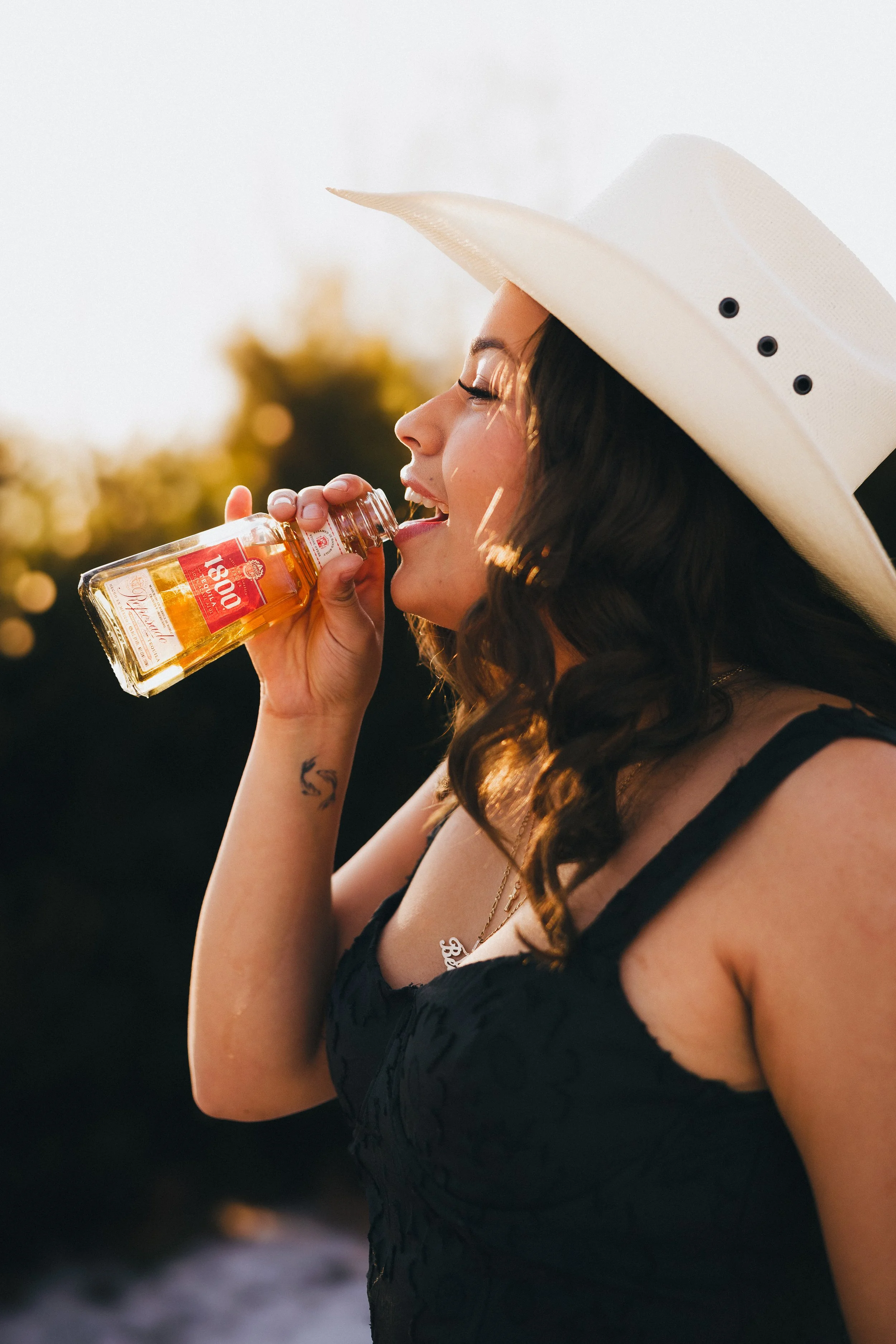 A woman wearing a white cowboy hat and a black dress is drinking from a small bottle of tequila outdoors during sunset.