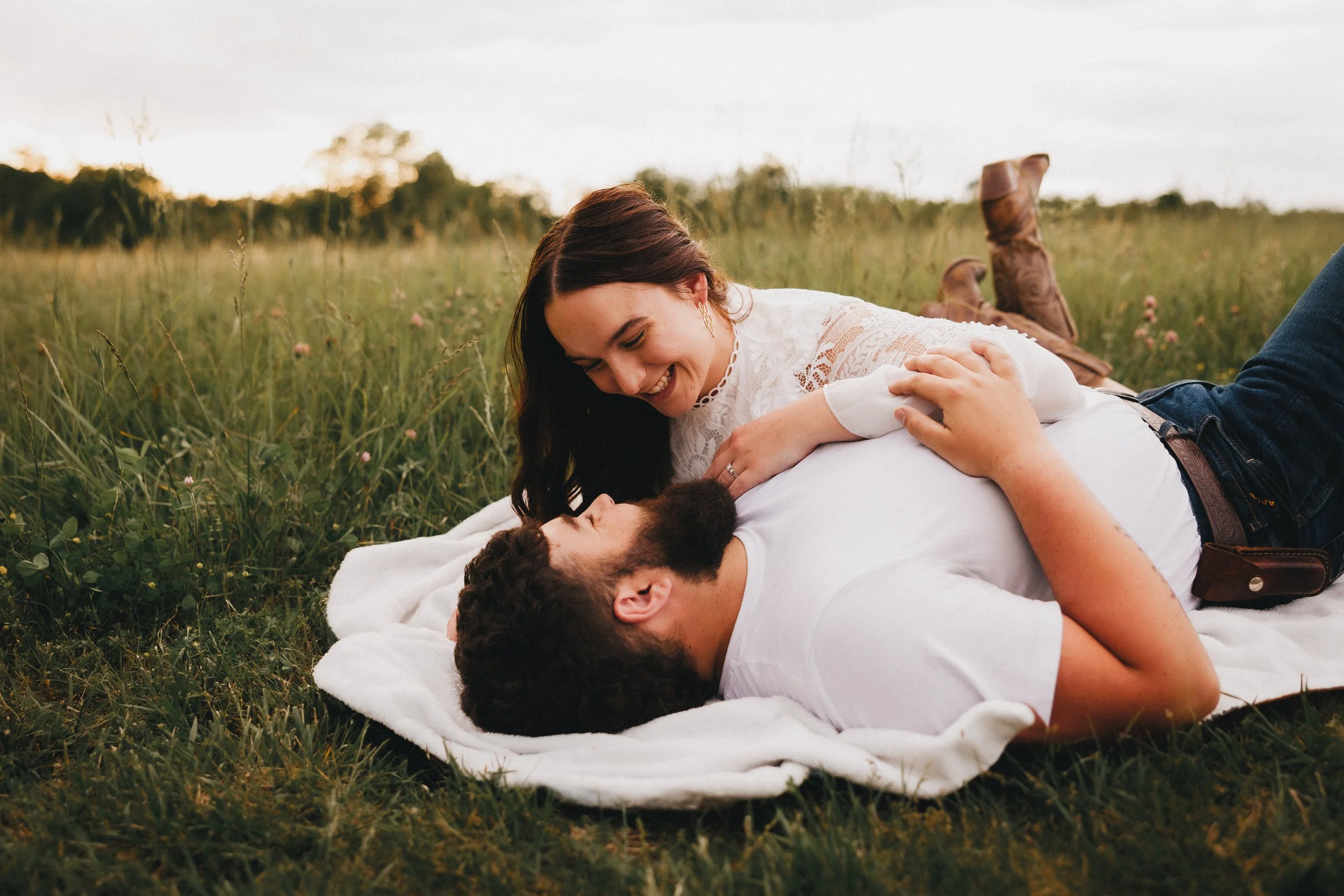 A couple lying on a blanket in a grassy field, smiling and gazing at each other, during sunset.