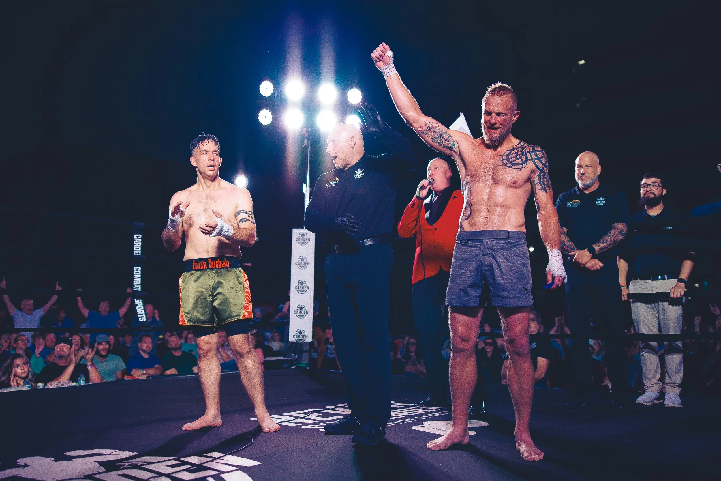 Two mixed martial arts fighters standing in a ring with a referee raising the hand of one fighter as the winner, surrounded by officials and an audience. Shot at Evergy Plaza in Topeka, Kansas. 