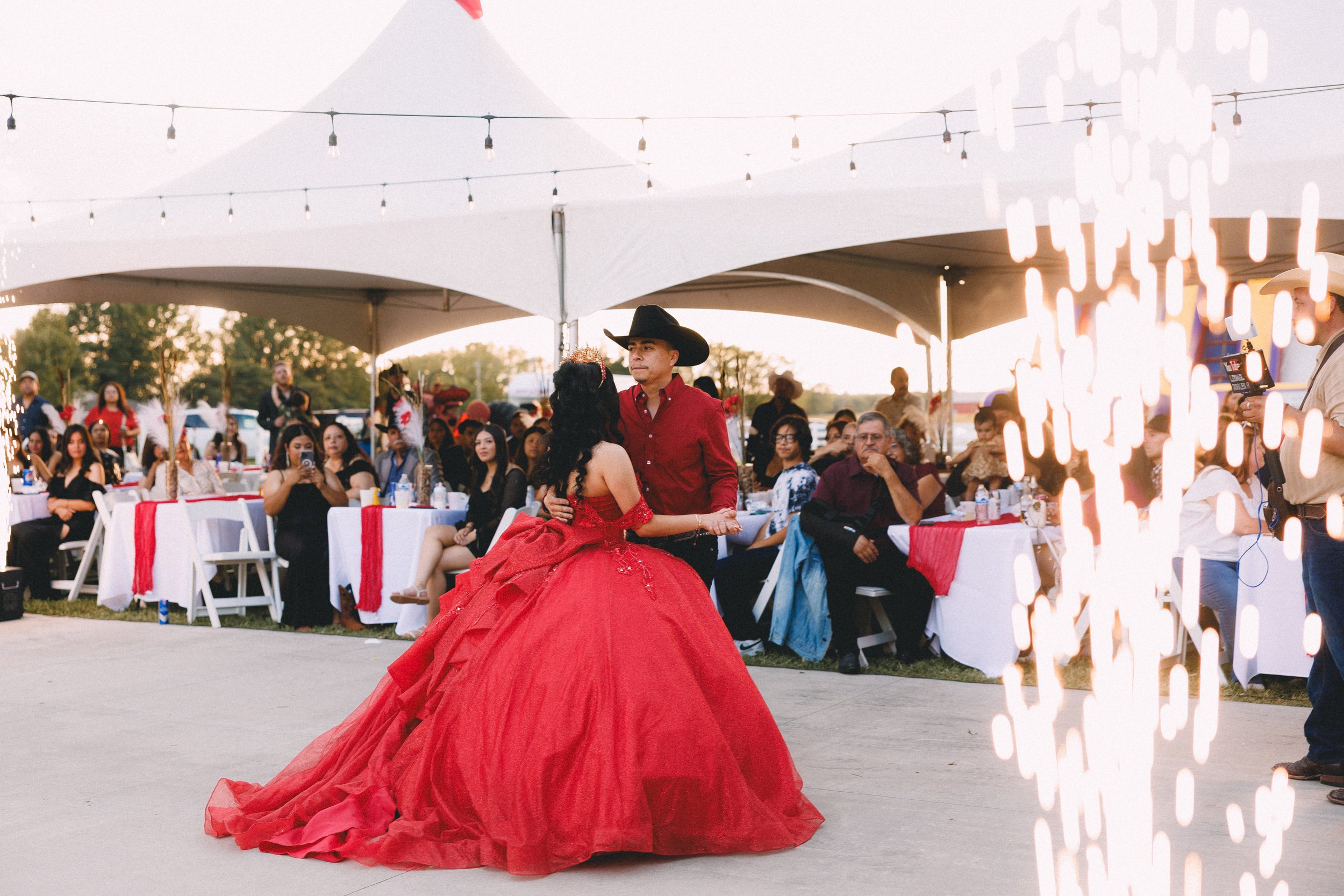 A couple wearing a red ball gown and cowboy hat dancing at an outdoor event under a large white tent, with guests seated at decorated tables watching.